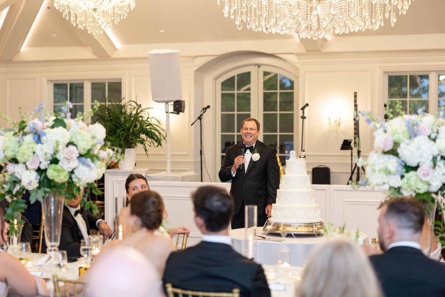 Man in tuxedo giving a speech at elegant wedding reception with floral arrangements and tiered cake.