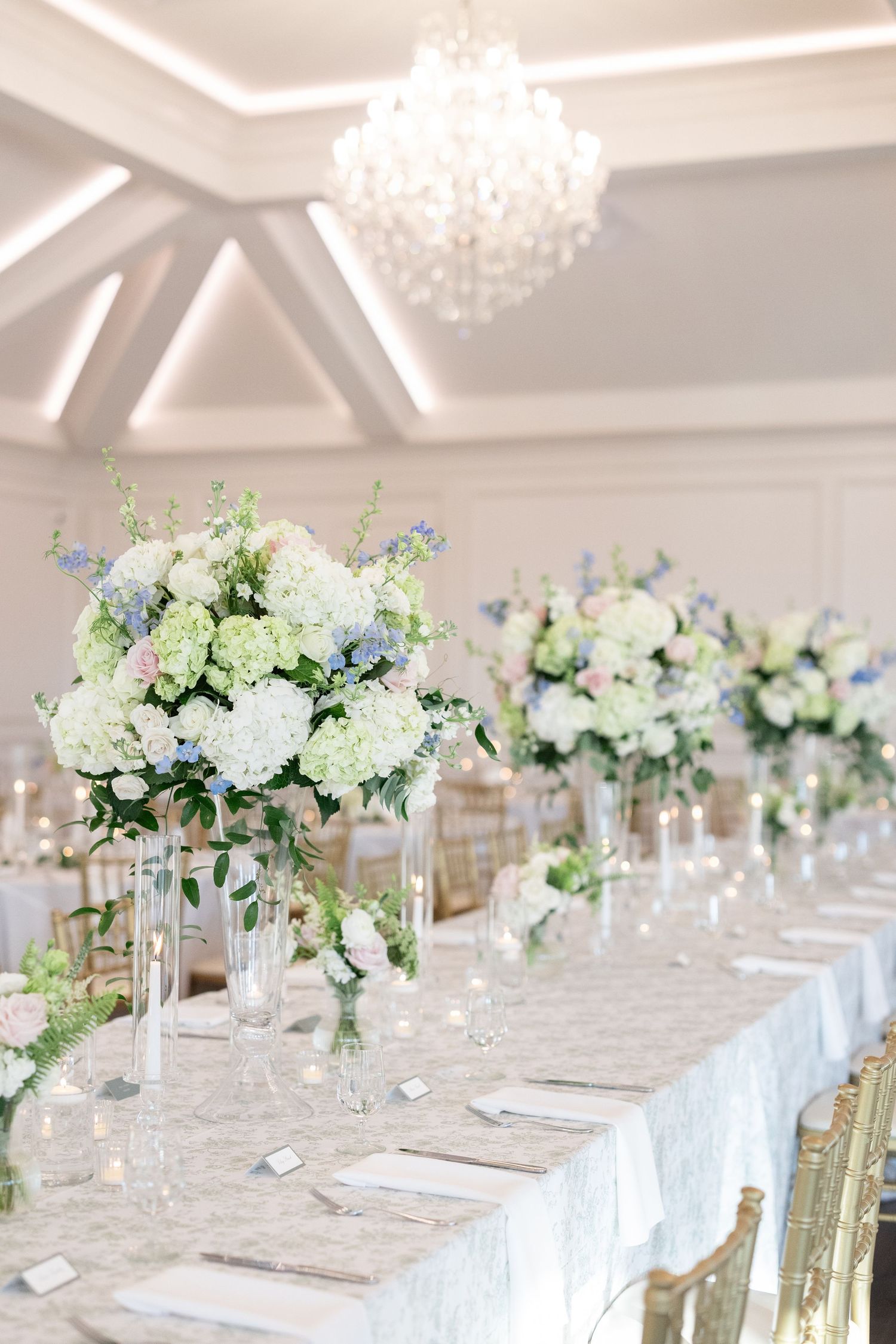 Elegant wedding reception table with tall white and blue hydrangea floral centerpieces and crystal chandelier.