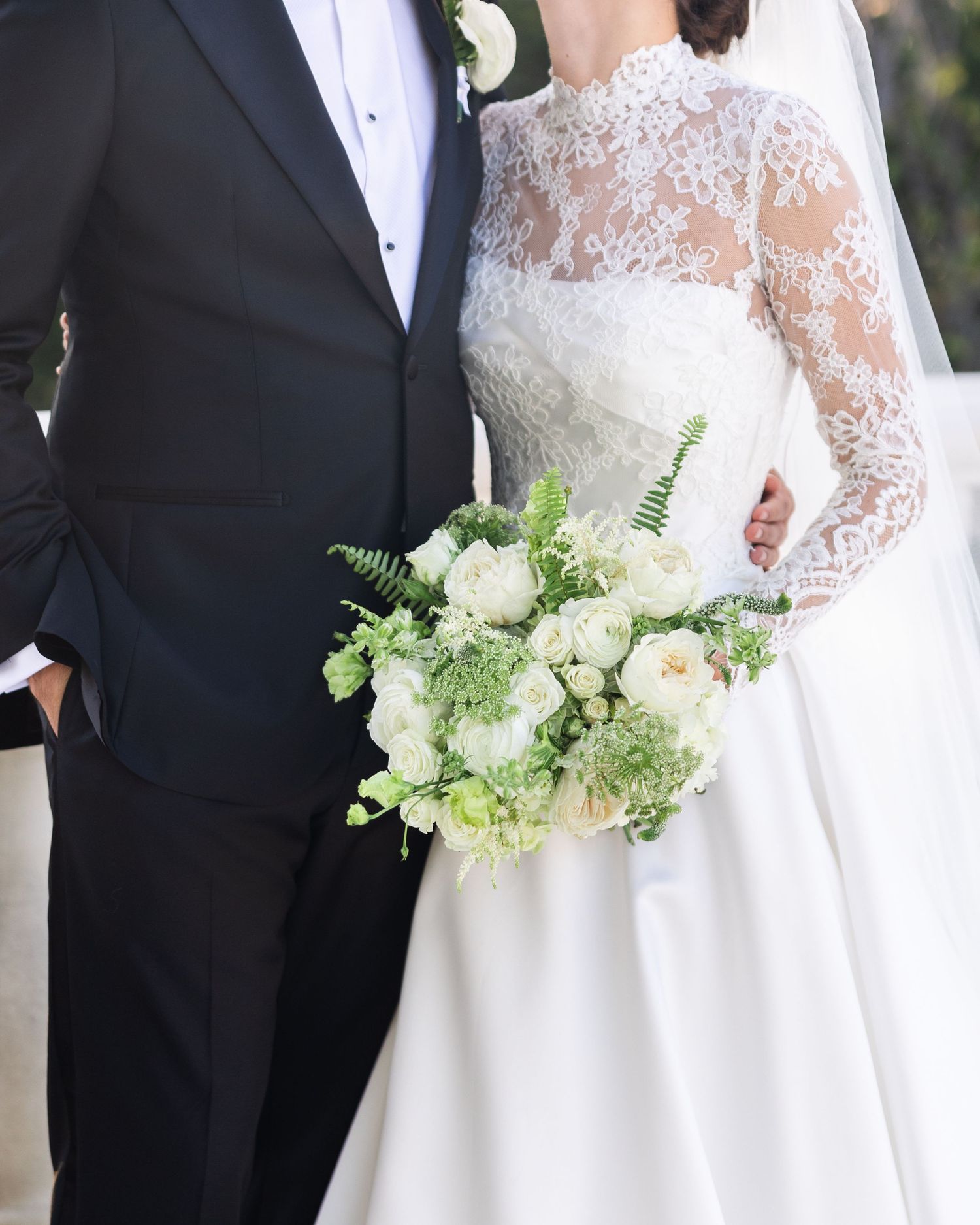 Close-up of bride in lace long-sleeve gown holding a lush white and green bouquet beside groom.