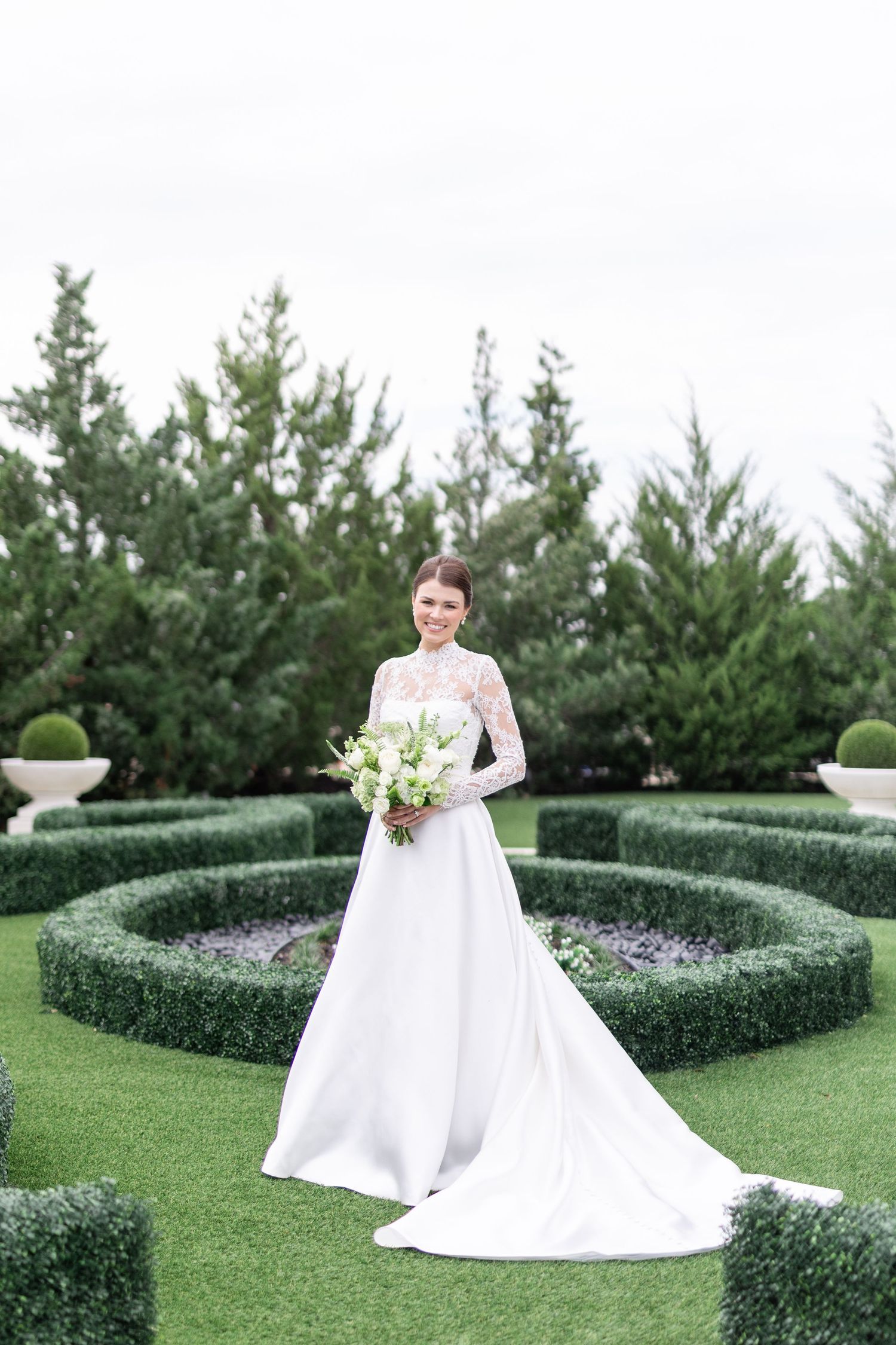 Bride in long-sleeve lace wedding gown with train holding white bouquet in formal garden.