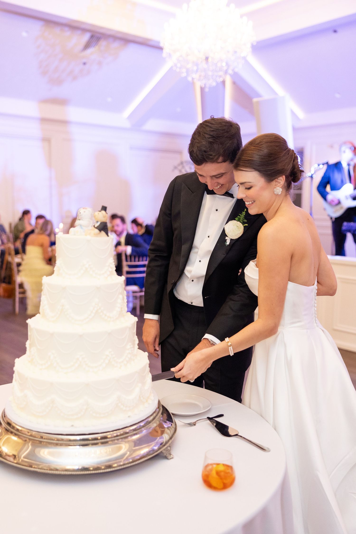 Bride and groom smiling while cutting four-tier white wedding cake at elegant reception.
