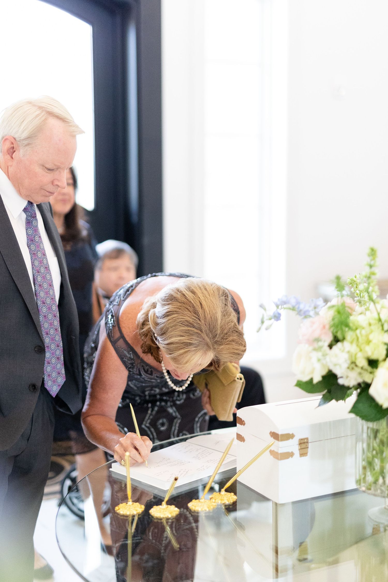 Wedding guest signing a guestbook at a glass table with floral arrangements at an elegant wedding venue.
