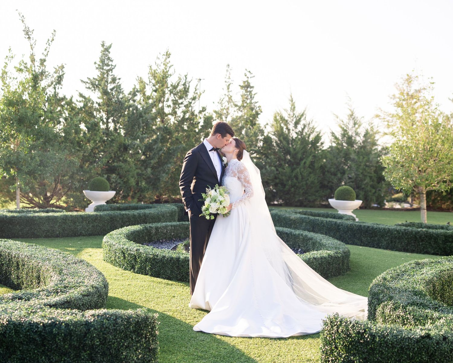 Bride and groom share a kiss in a manicured garden maze at golden hour.