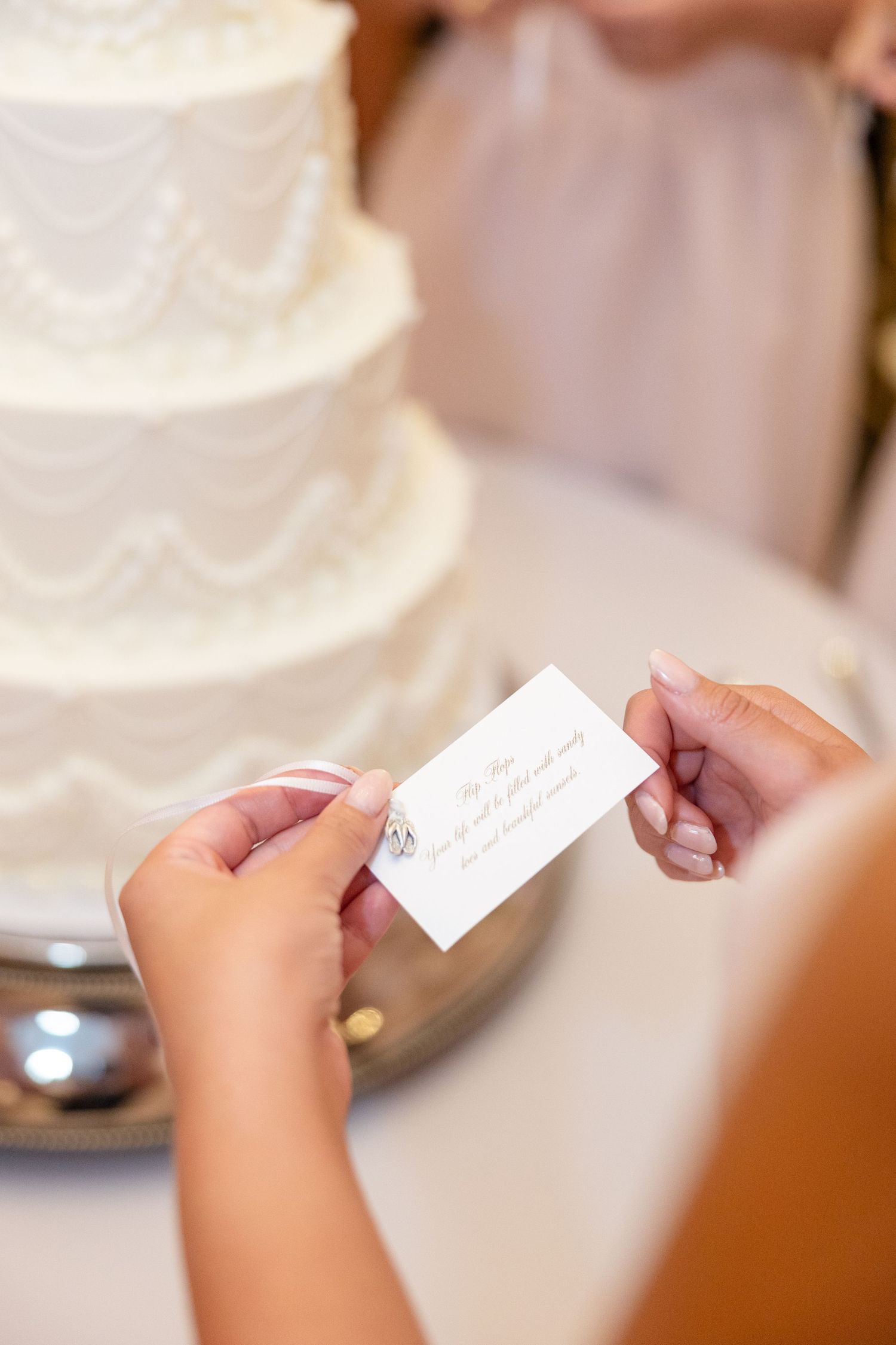 Hands holding small wedding card in front of elegant white tiered wedding cake.