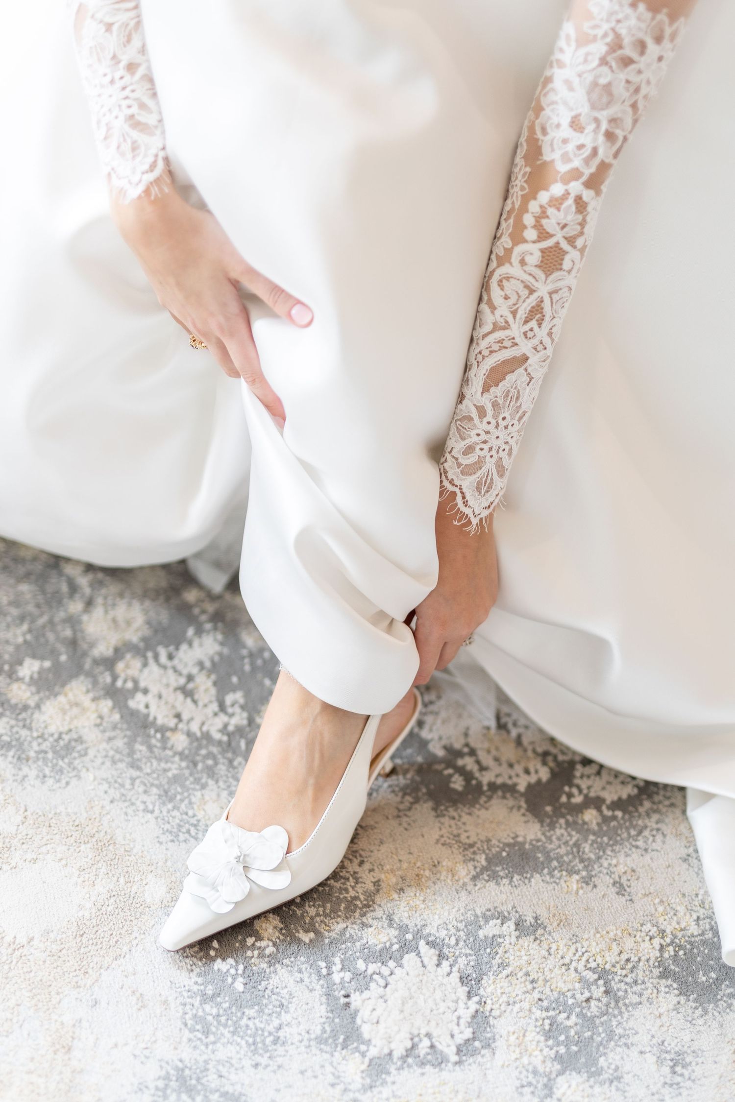 Close-up of bride slipping on white floral pointed-toe wedding heels with lace dress.