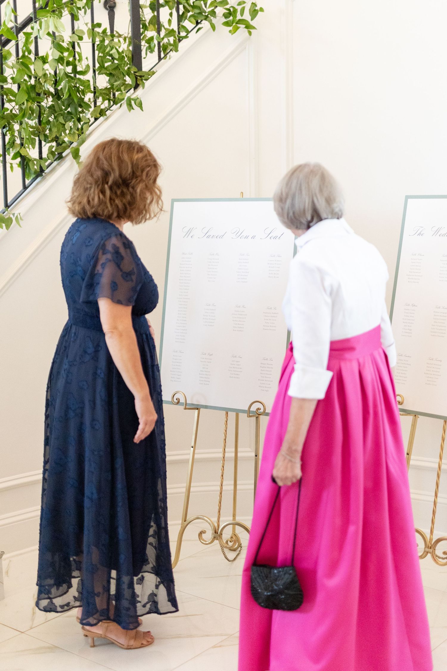 Two elegantly dressed women reviewing a seating chart display on a gold easel at a formal wedding reception.