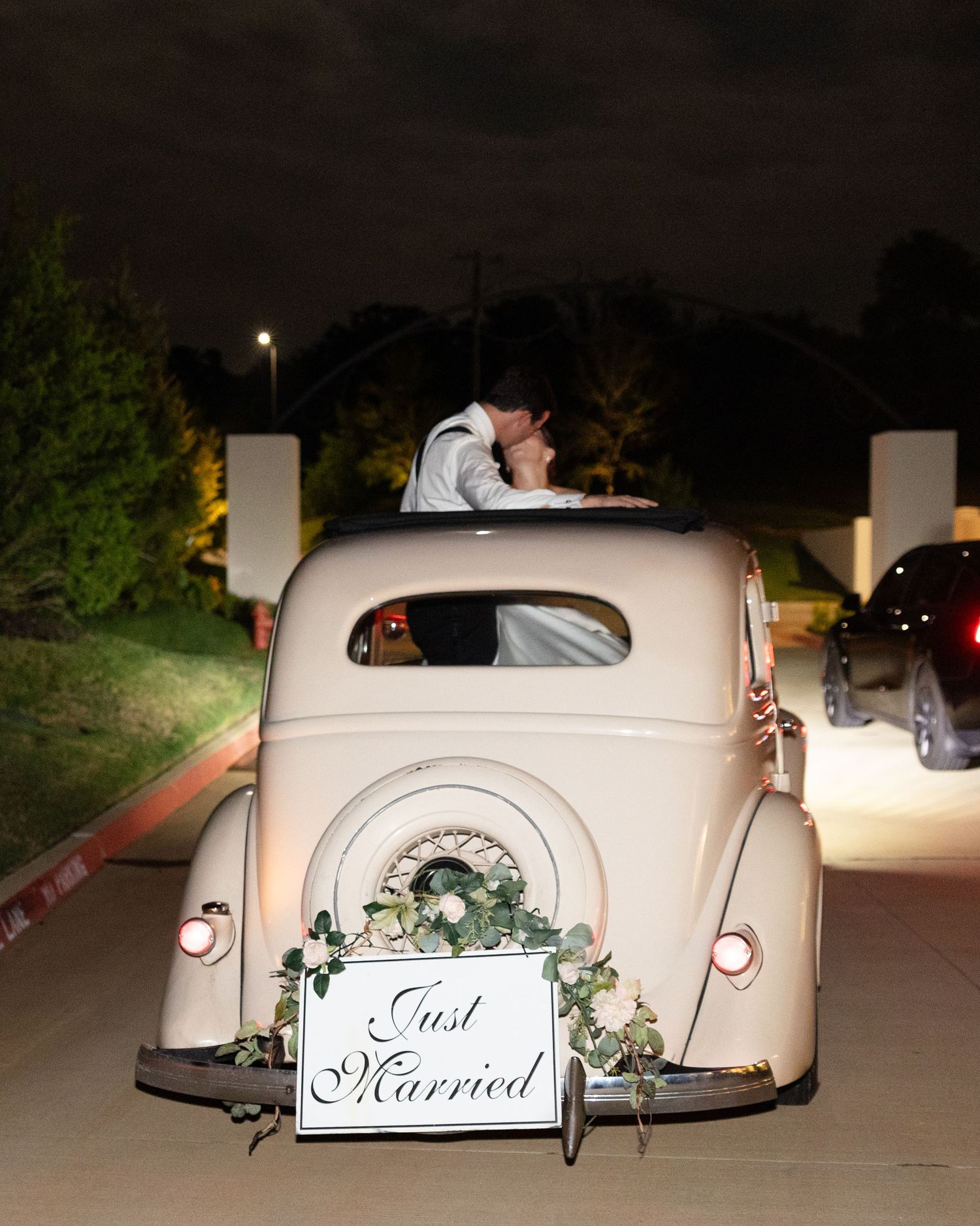 Vintage cream getaway car with Just Married sign and floral wreath departs wedding at night.