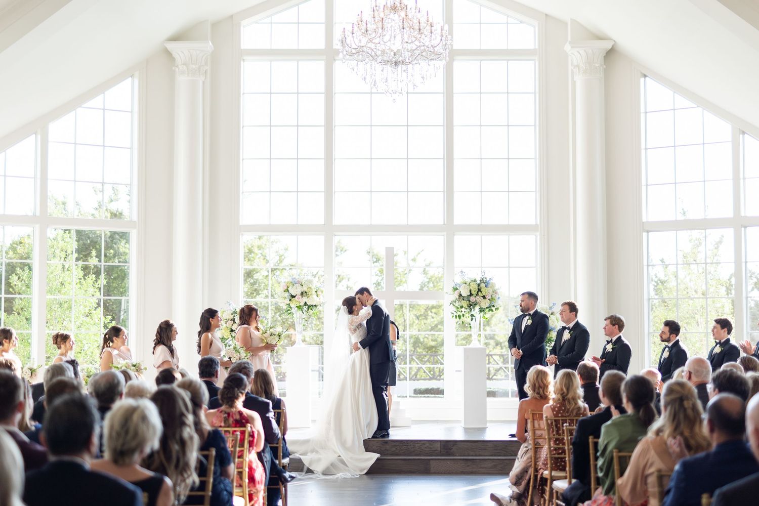 Elegant wedding ceremony in a bright chapel with chandelier, guests seated, and couple at the altar.
