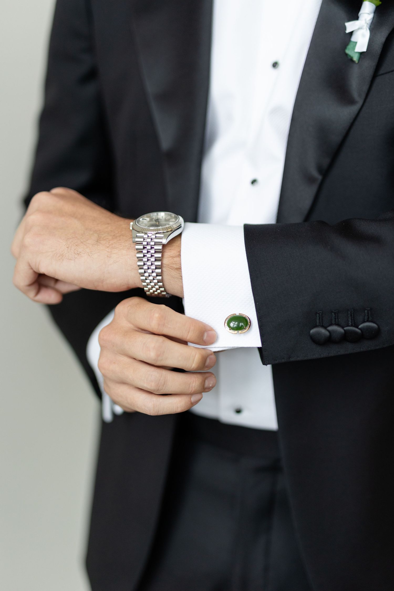 Groom adjusting green cufflinks on white dress shirt while wearing a silver watch and black tuxedo.