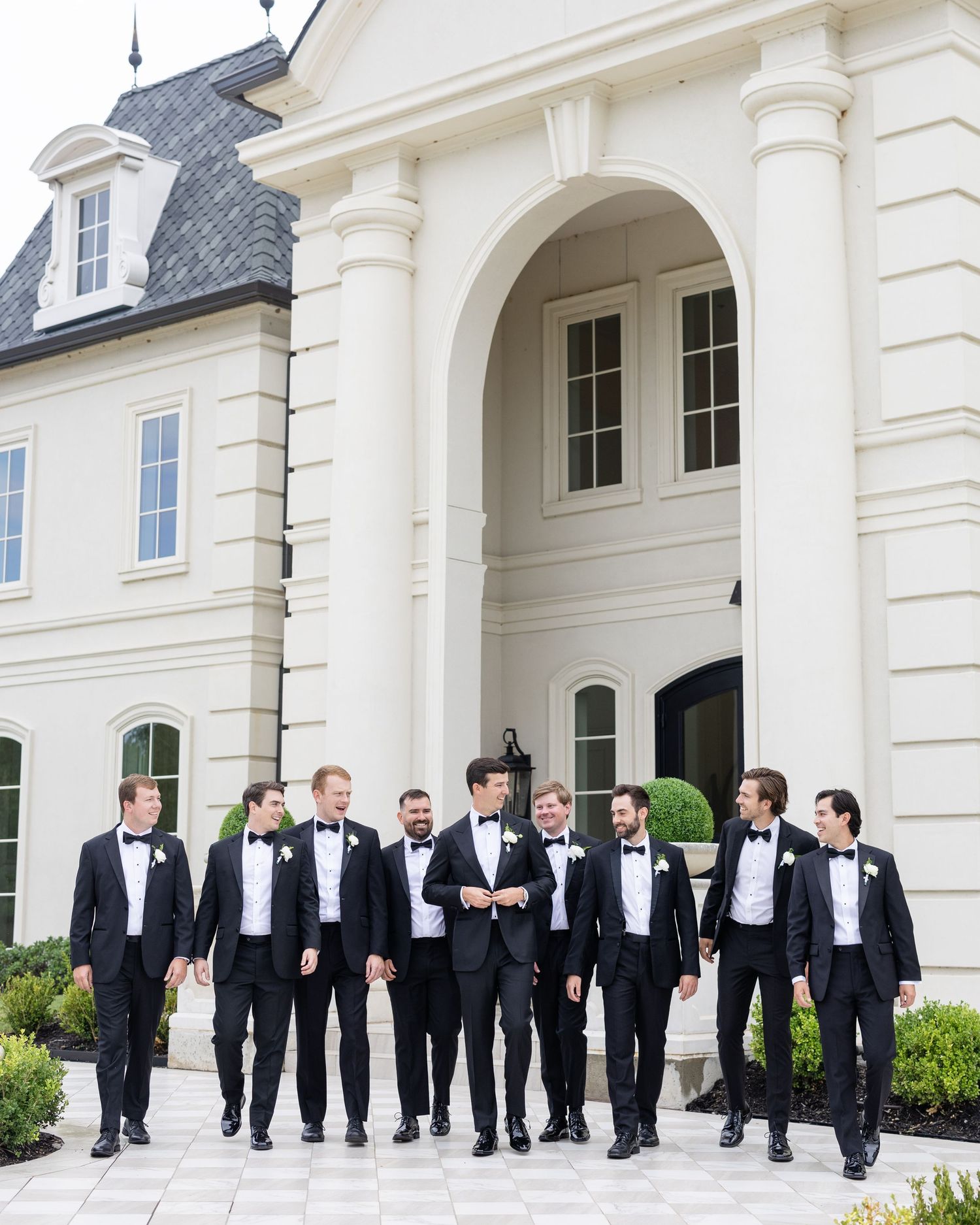 Groomsmen in classic black tuxedos walk together outside a grand white estate wedding venue.