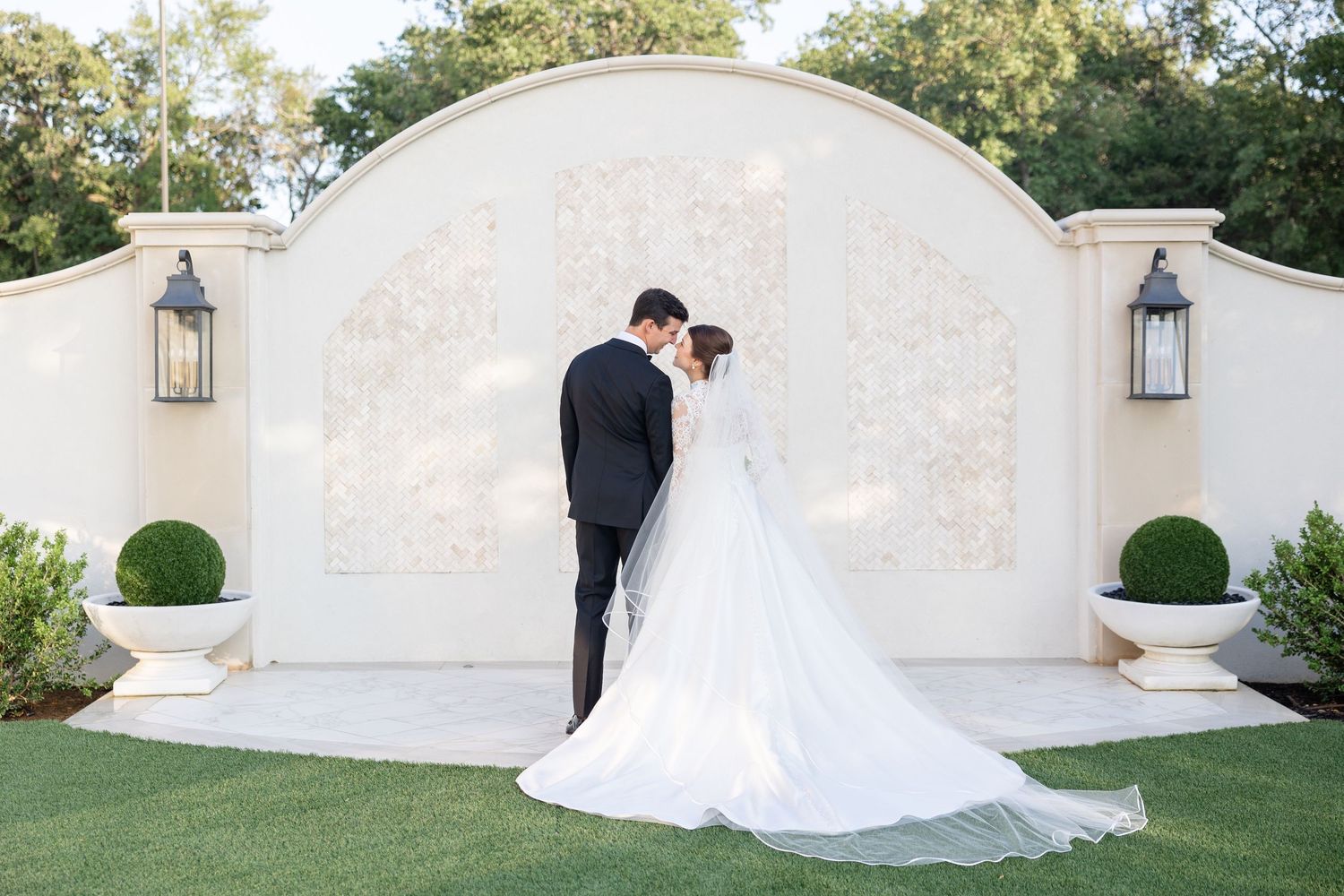 Bride and groom share a kiss before a decorative white arch flanked by lanterns in a garden.
