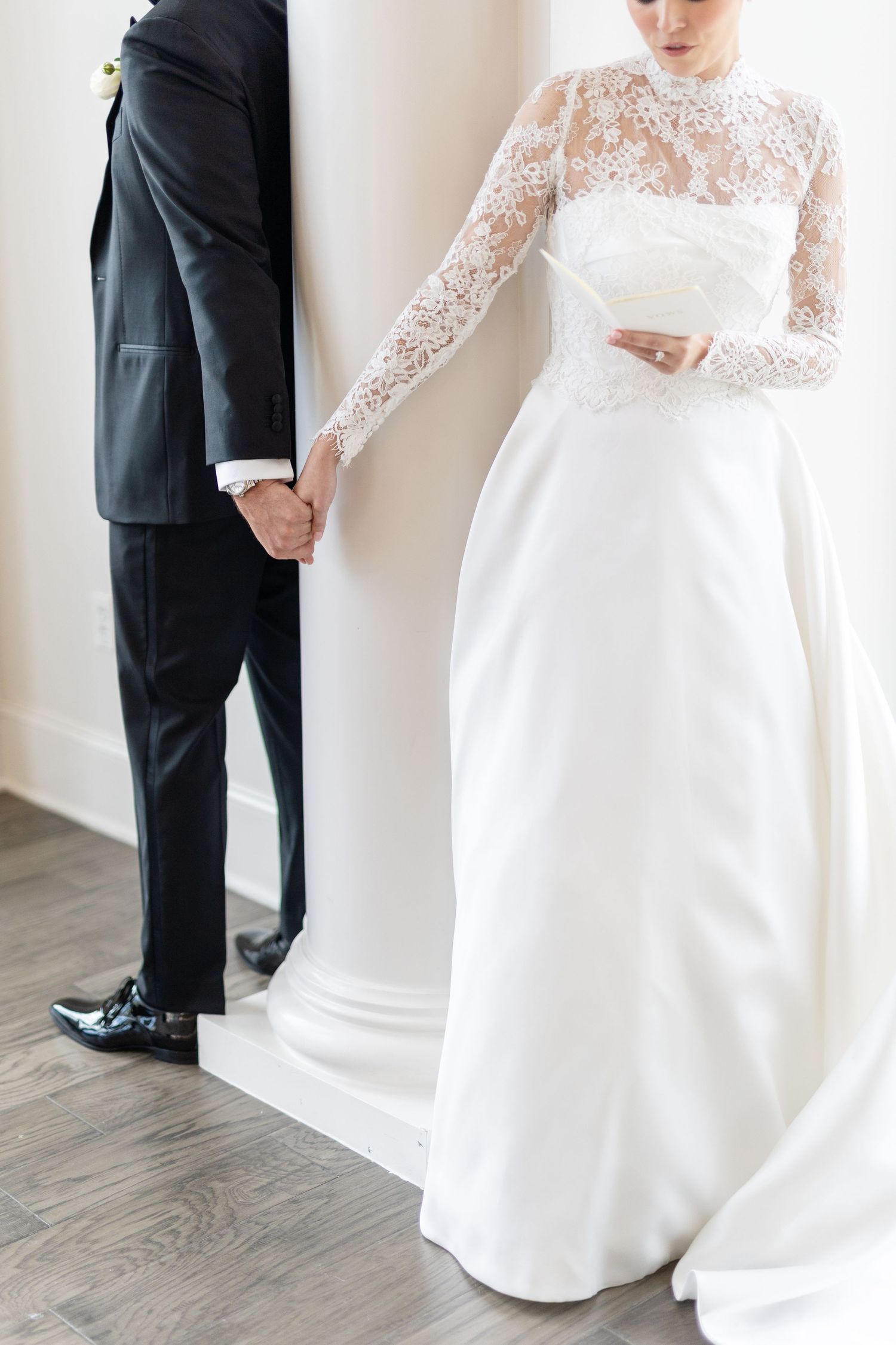 Bride in high-neck lace long-sleeve wedding gown holding groom's hand beside white column.