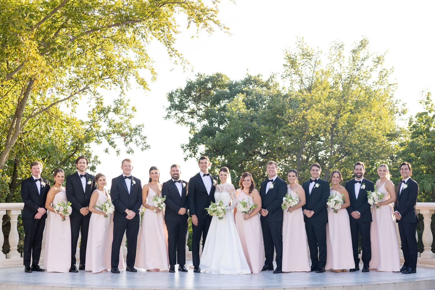 Wedding party in blush bridesmaid dresses and black tuxedos posing outdoors on a terrace with greenery.