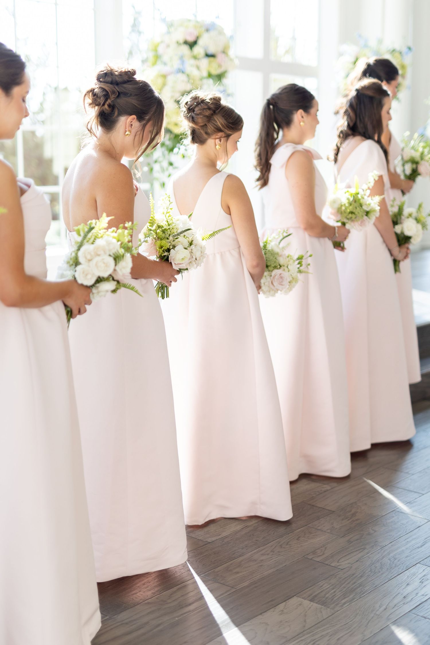 Bridesmaids in blush pink gowns holding white floral bouquets standing in a row at a wedding ceremony.