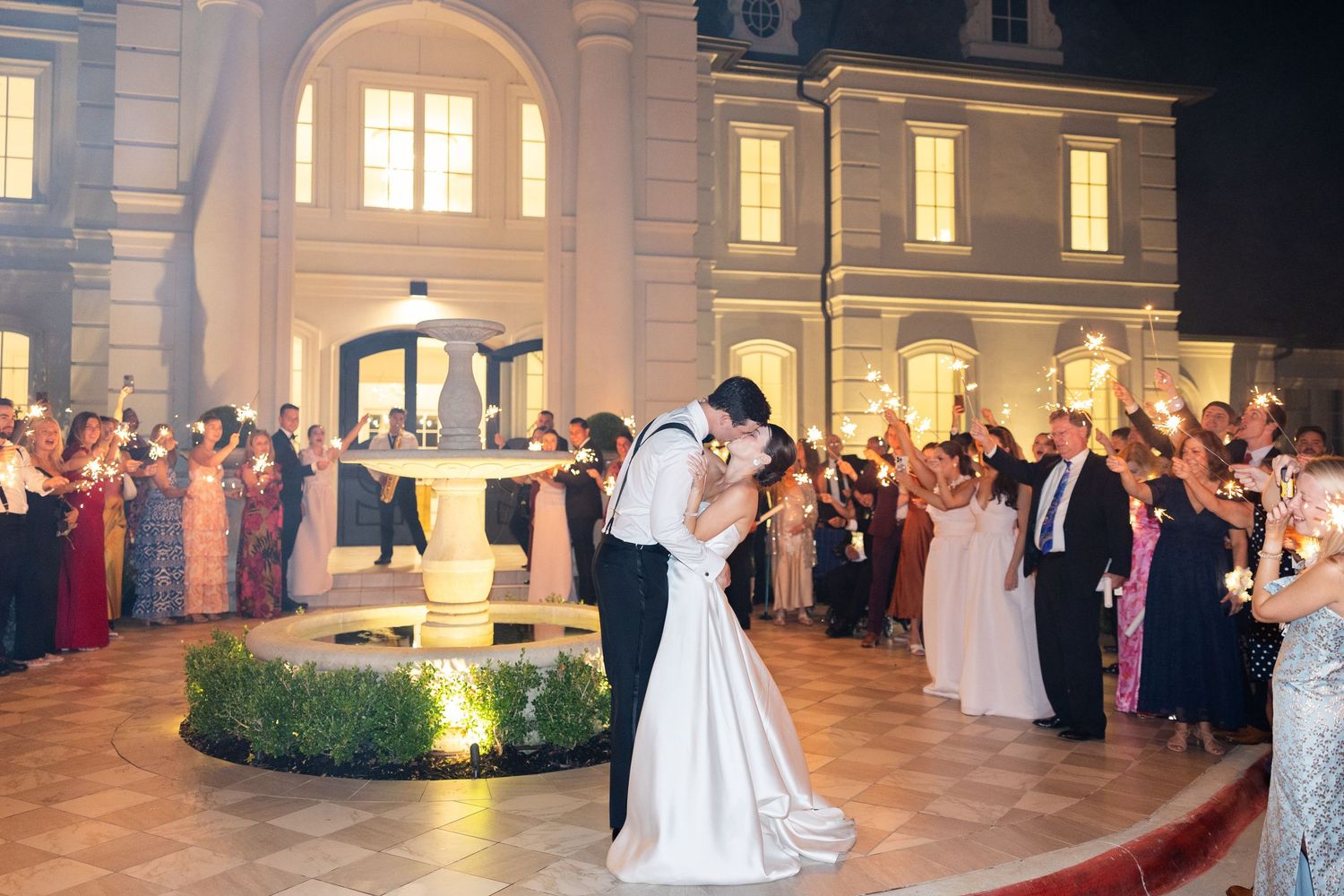 Newlyweds kiss near fountain as guests wave sparklers during outdoor wedding sendoff.