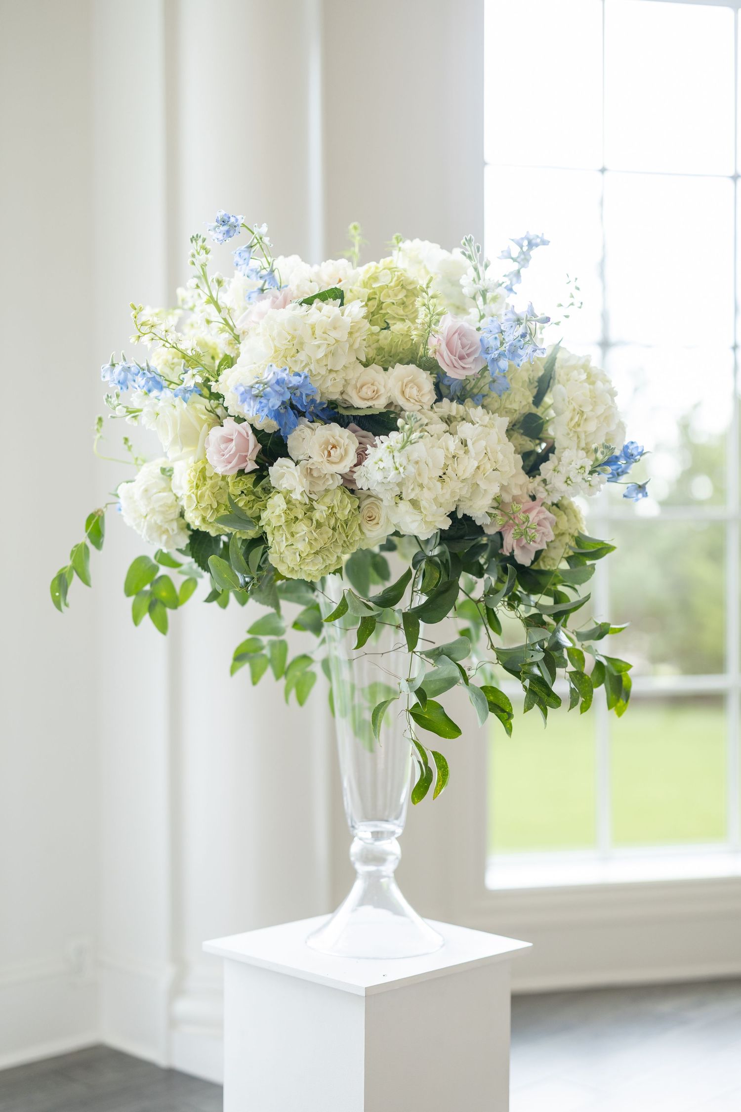 Tall glass vase arrangement of white hydrangeas, blue delphiniums, and pink roses on white pedestal.