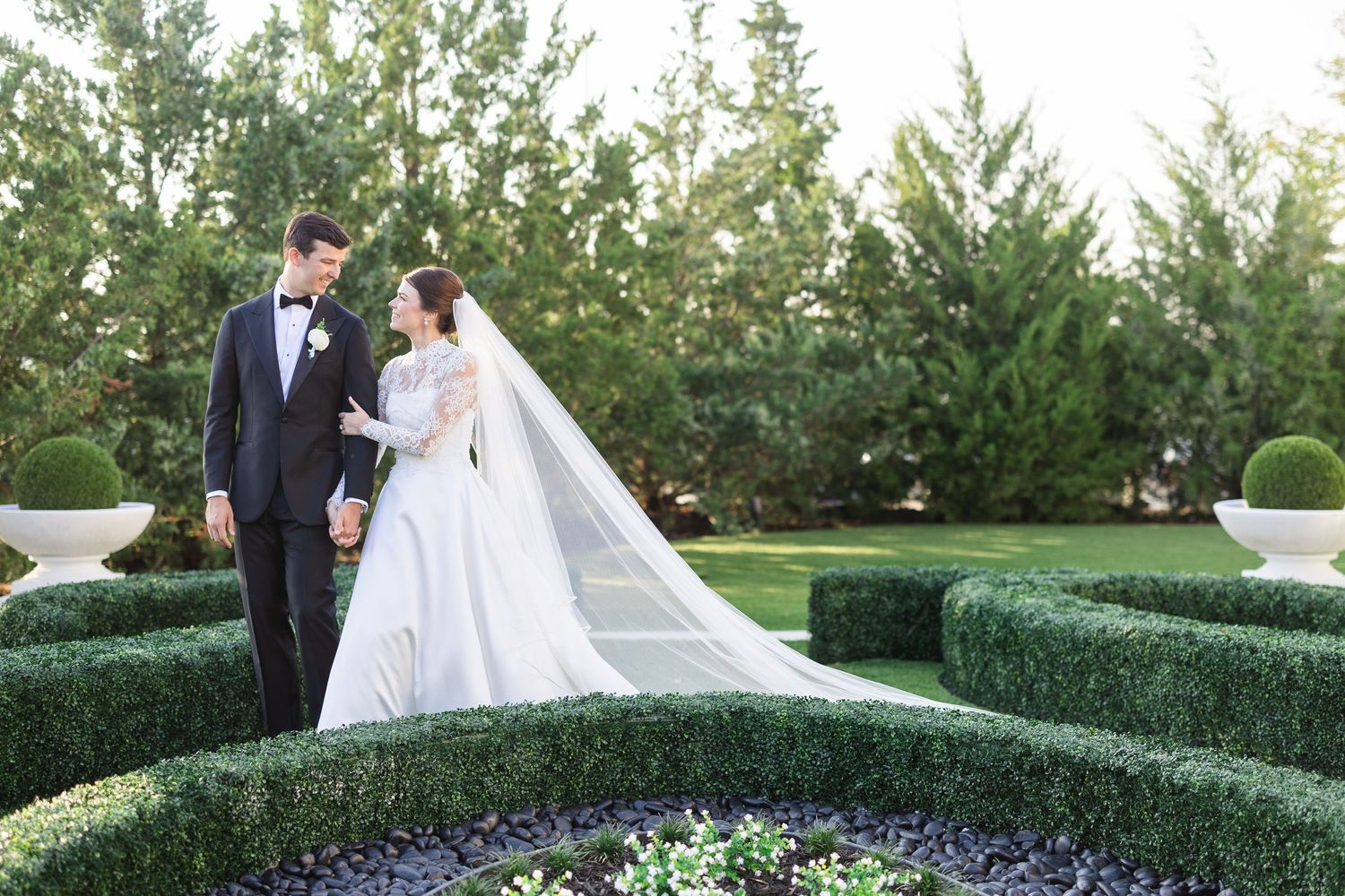 Bride and groom holding hands in a lush garden maze with manicured hedges, bride wearing a long veil.