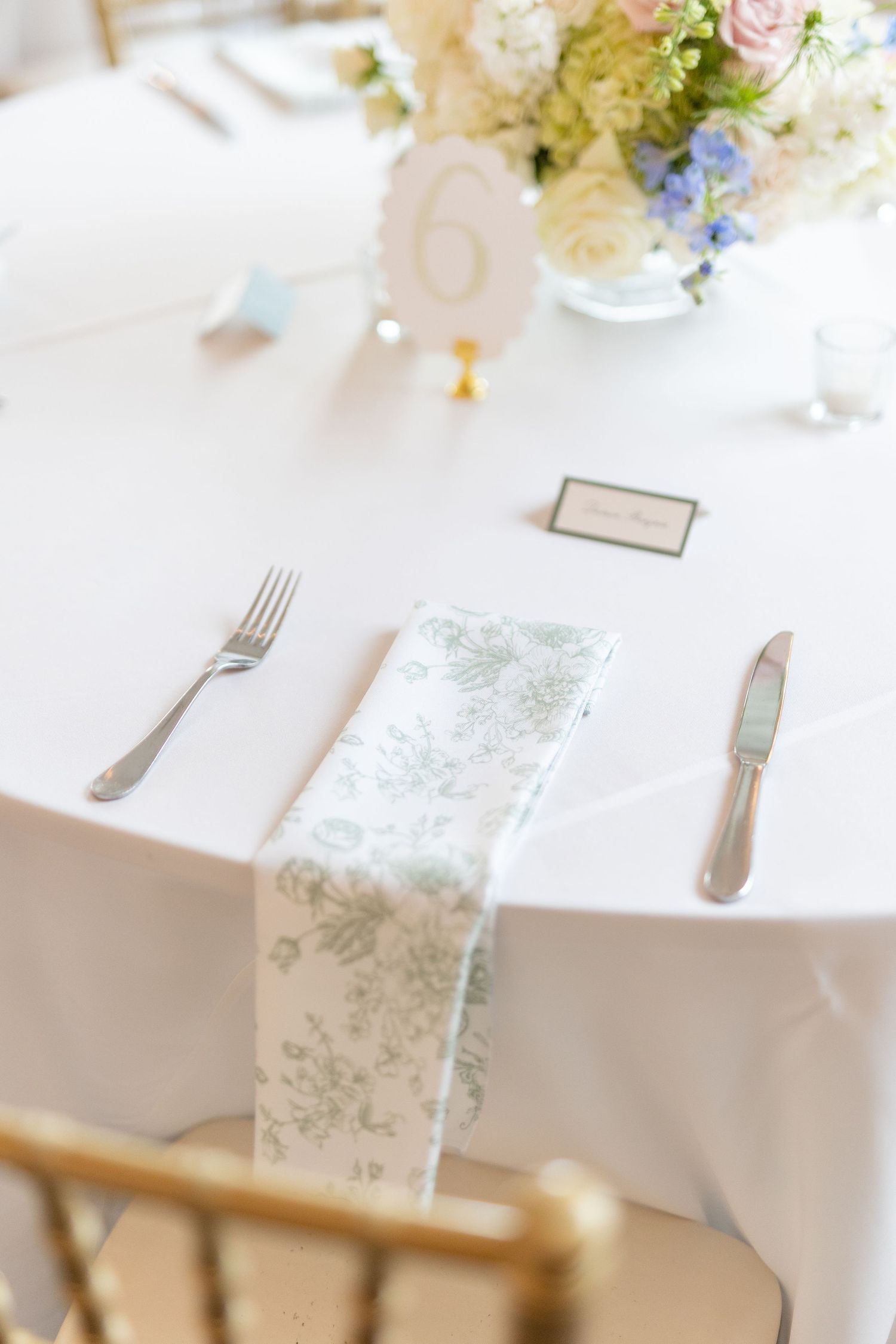 Elegant wedding table setting with floral napkin, silver cutlery, place card, and pastel centerpiece.