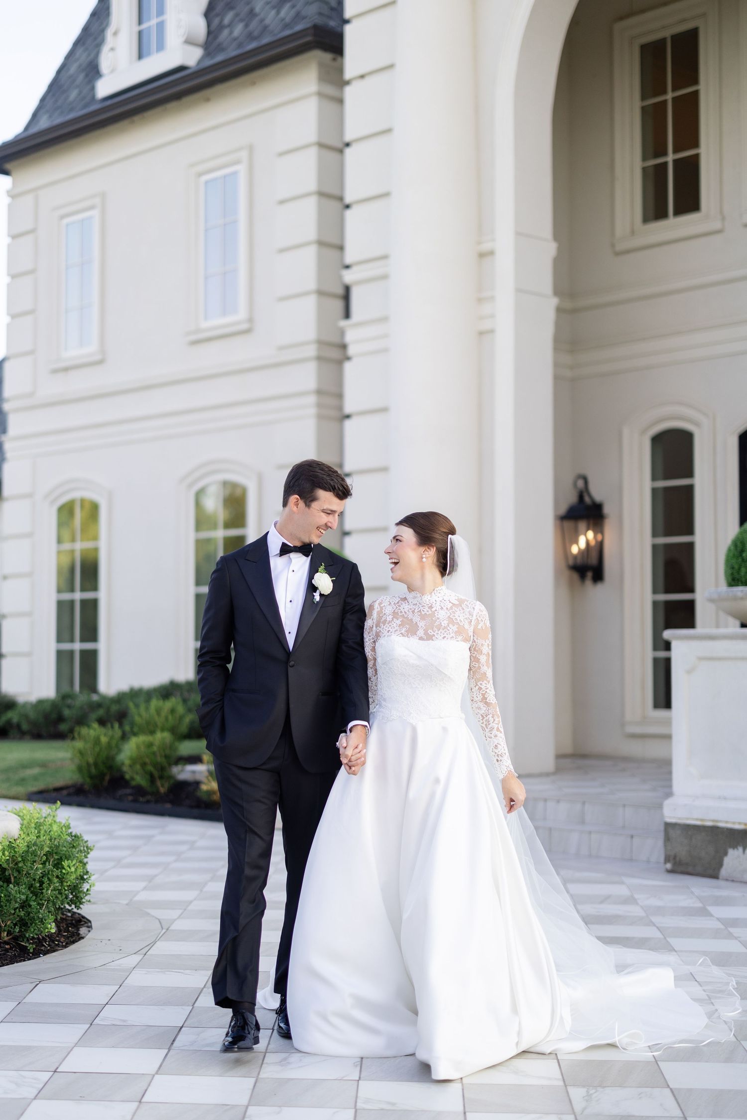 Smiling bride and groom walking hand in hand outside a stunning white French chateau wedding venue.
