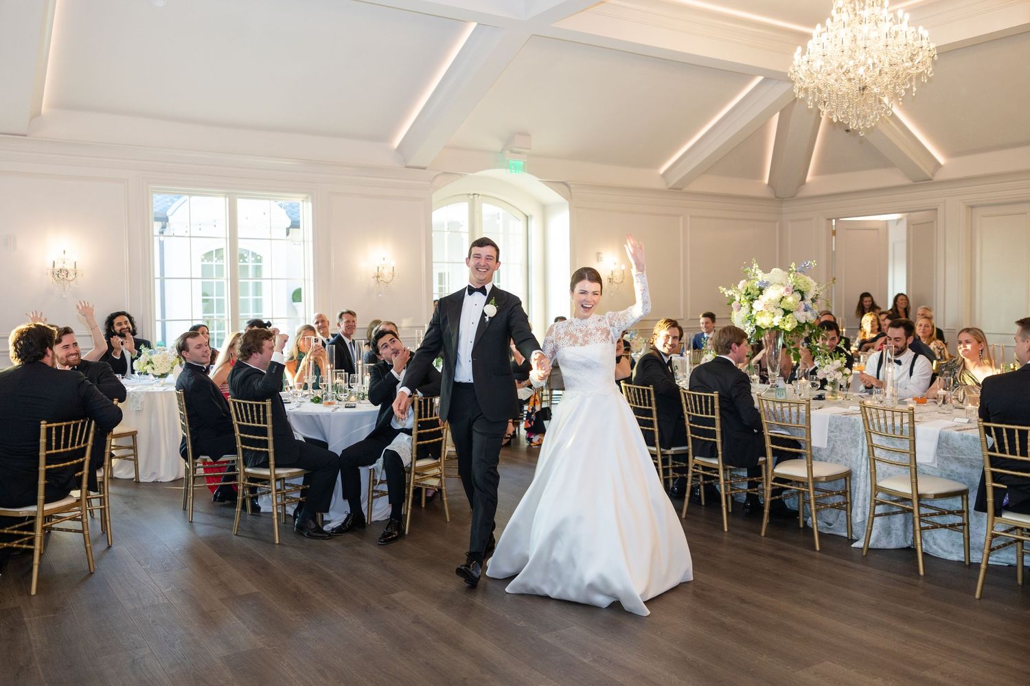 Bride and groom make grand entrance into elegant white wedding reception hall with guests seated at decorated tables.