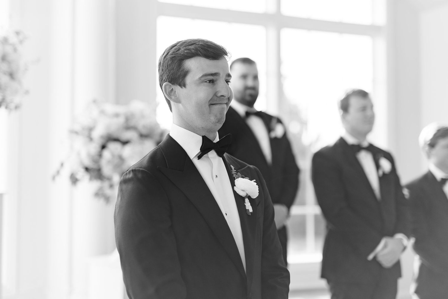 Black and white portrait of an emotional groom in a tuxedo watching his bride walk down the aisle.