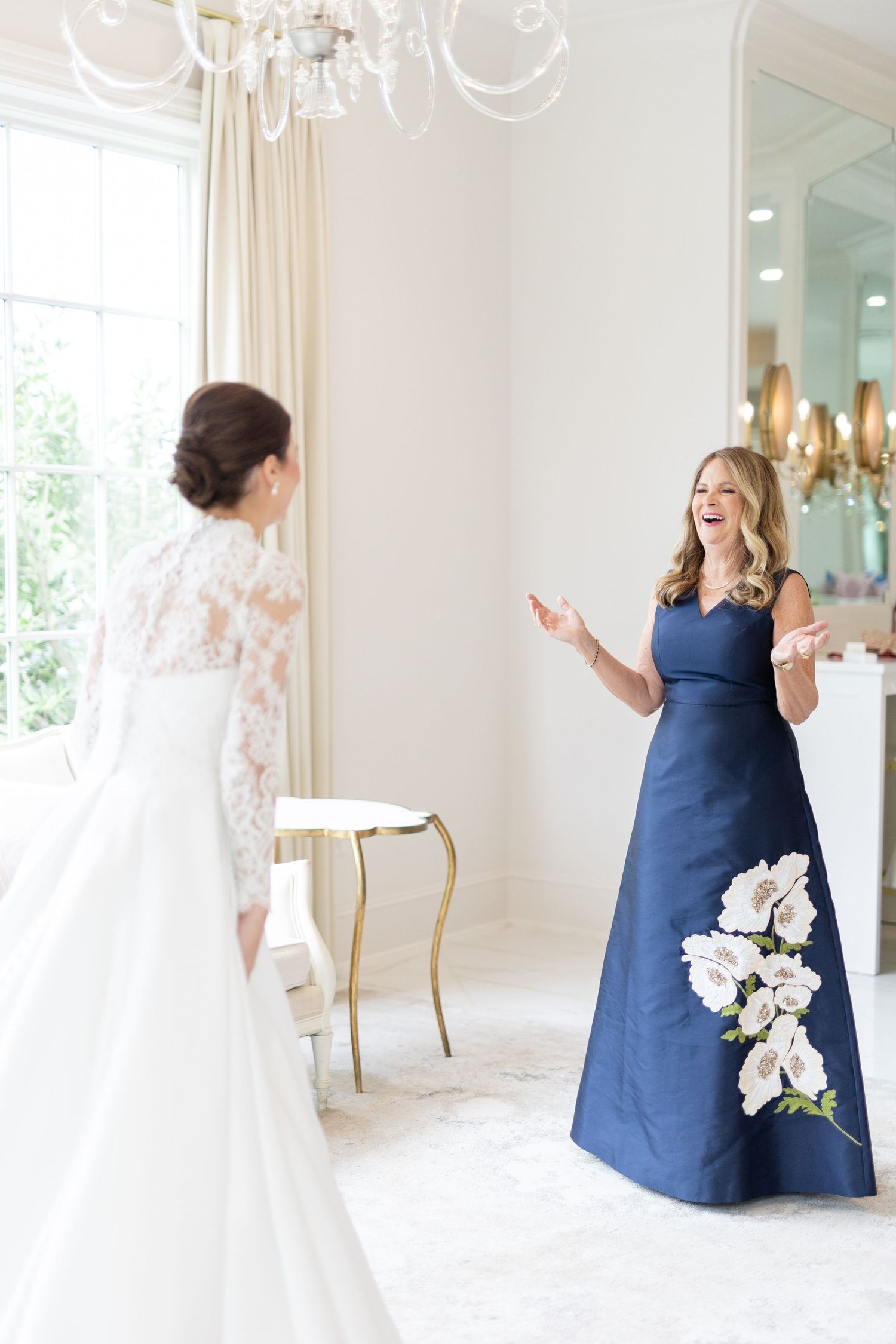 Woman in navy blue floral gown reacting with joy seeing bride in lace wedding dress.