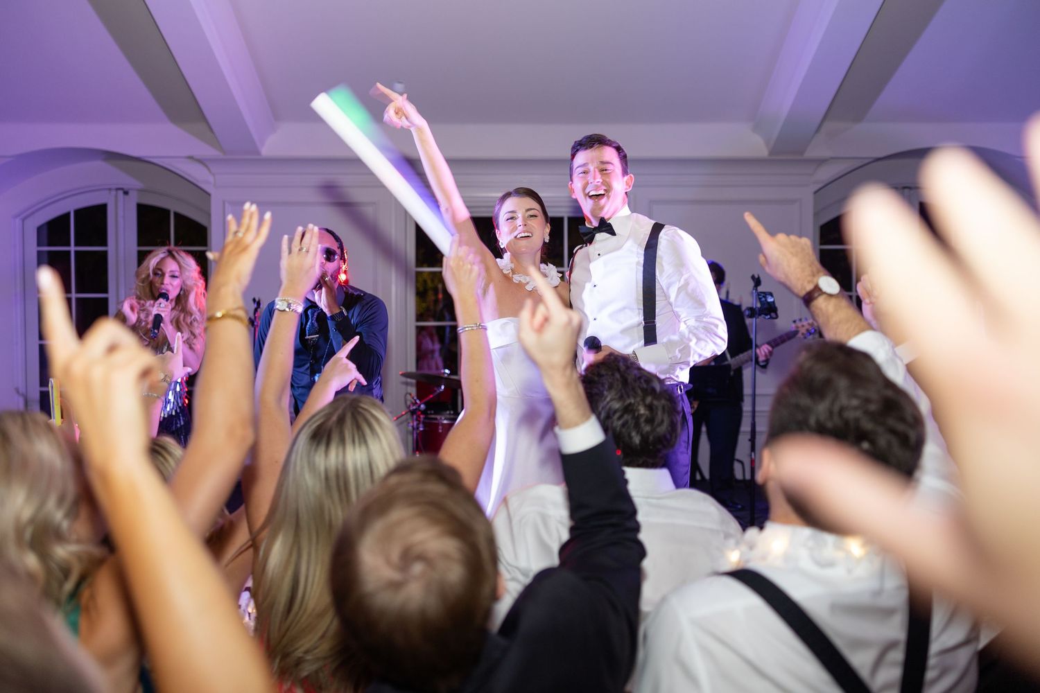 Bride and groom celebrate on dance floor as guests cheer with raised hands at wedding reception.