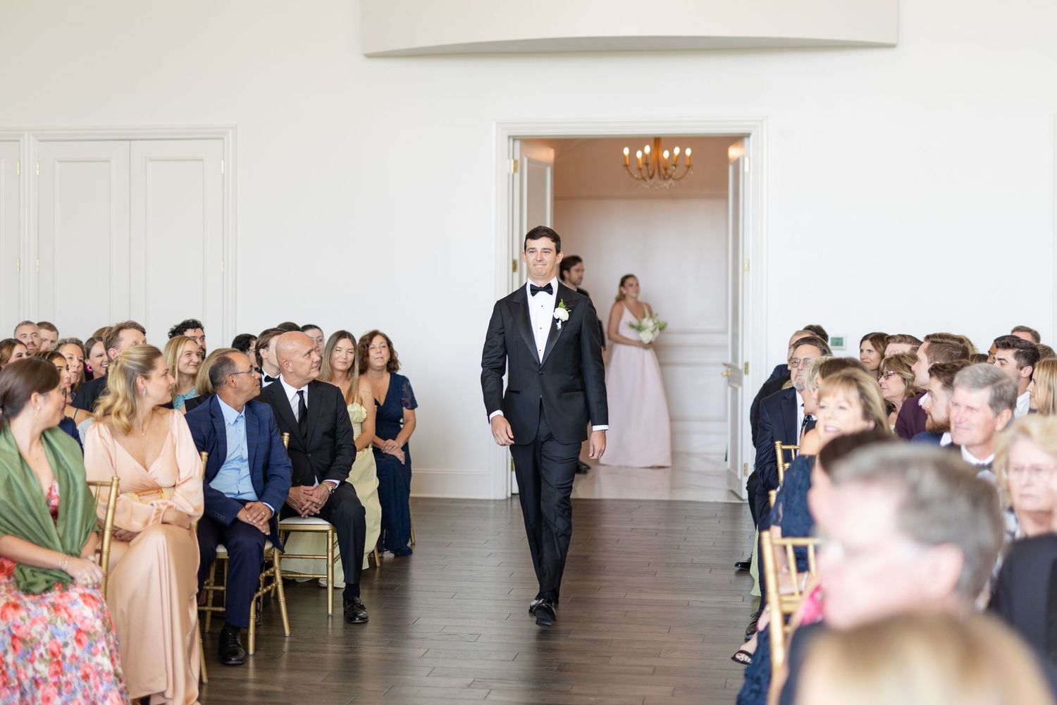 Groom in black tuxedo walking down the aisle as guests watch at an elegant white wedding venue.