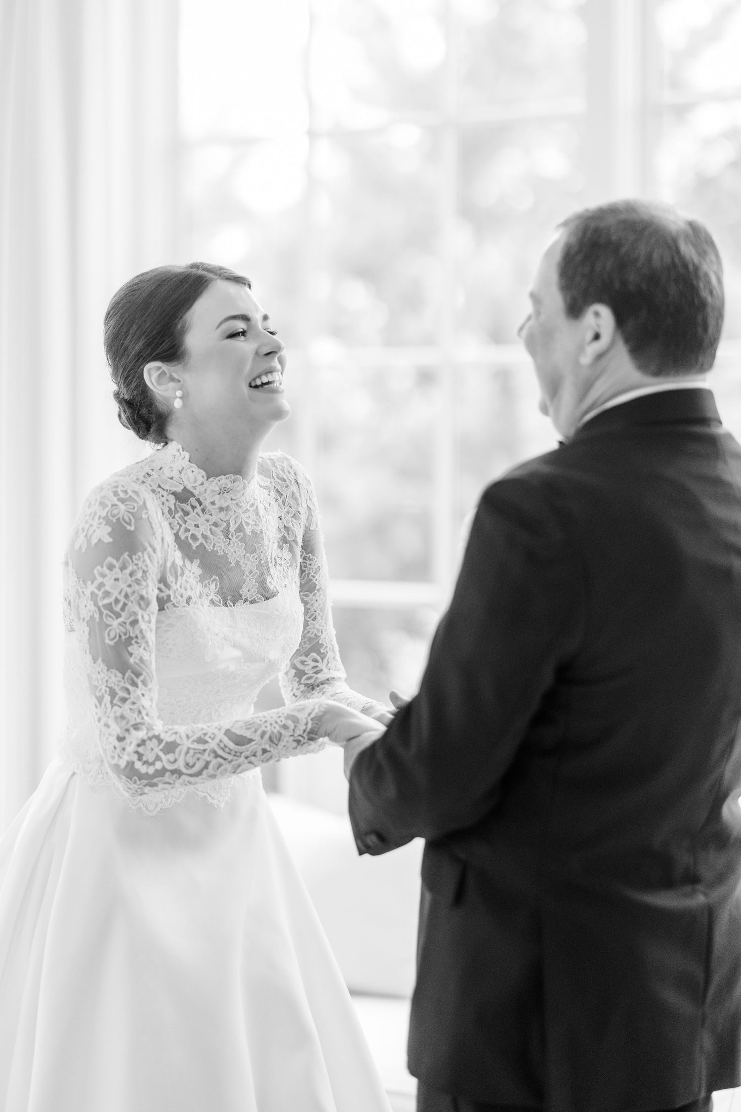 Bride in lace long-sleeve wedding gown laughing joyfully with man in suit during first look.