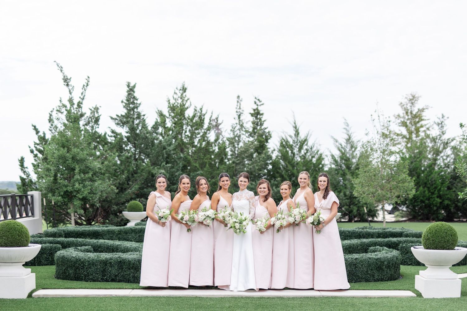 Bridesmaids in blush pink gowns stand with bride in white before a manicured garden backdrop.