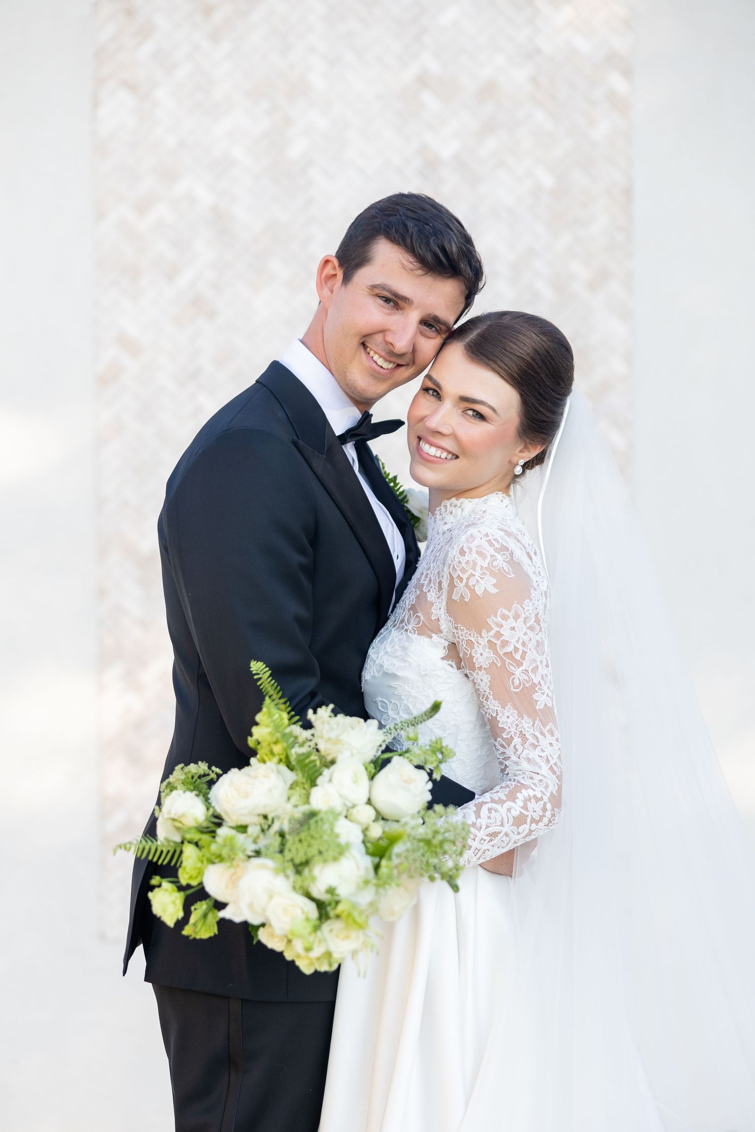 Joyful bride in long-sleeve lace gown and groom embrace, smiling before an elegant white backdrop.