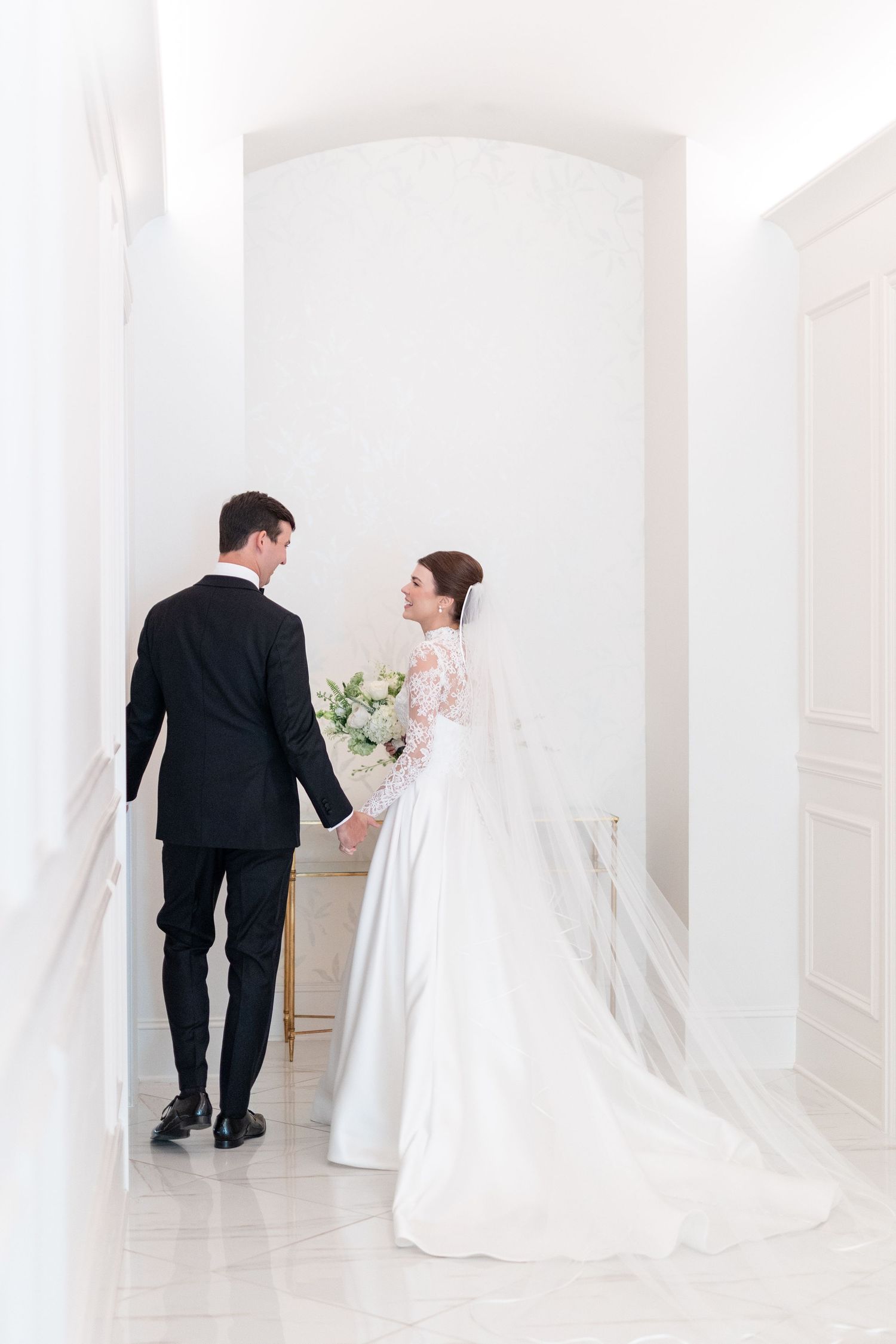 Bride in a lace wedding gown with long veil holding bouquet walks hand-in-hand with groom in white hallway.
