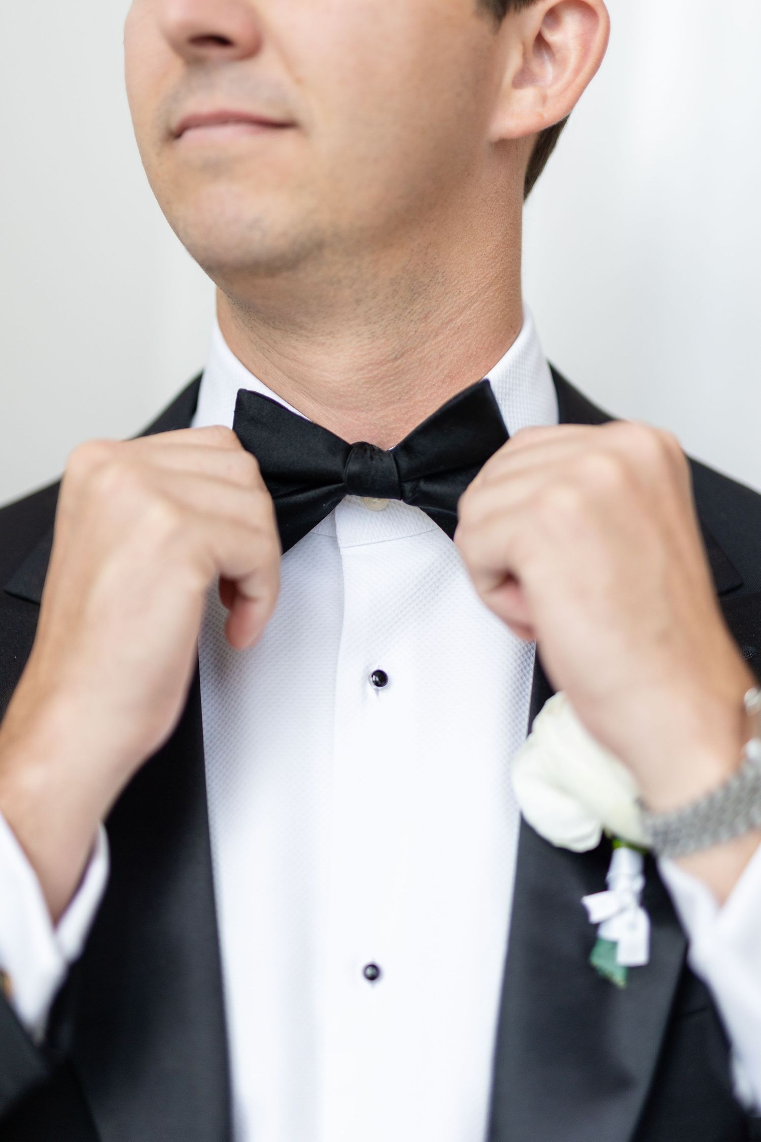Groom in black tuxedo adjusting bow tie with white boutonniere on lapel.