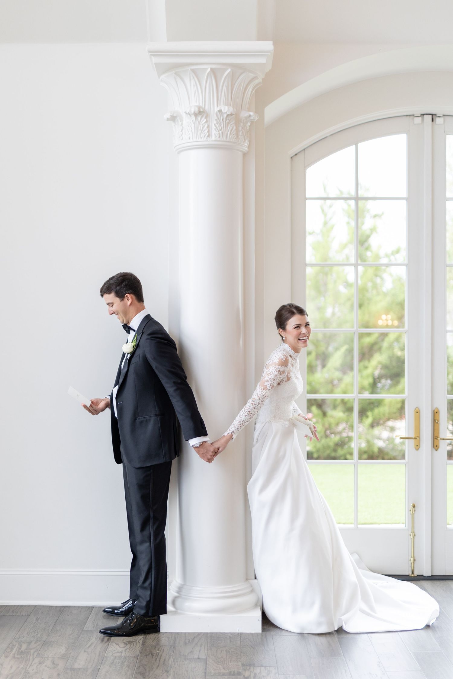 Bride and groom holding hands around white column reading vows before wedding ceremony.