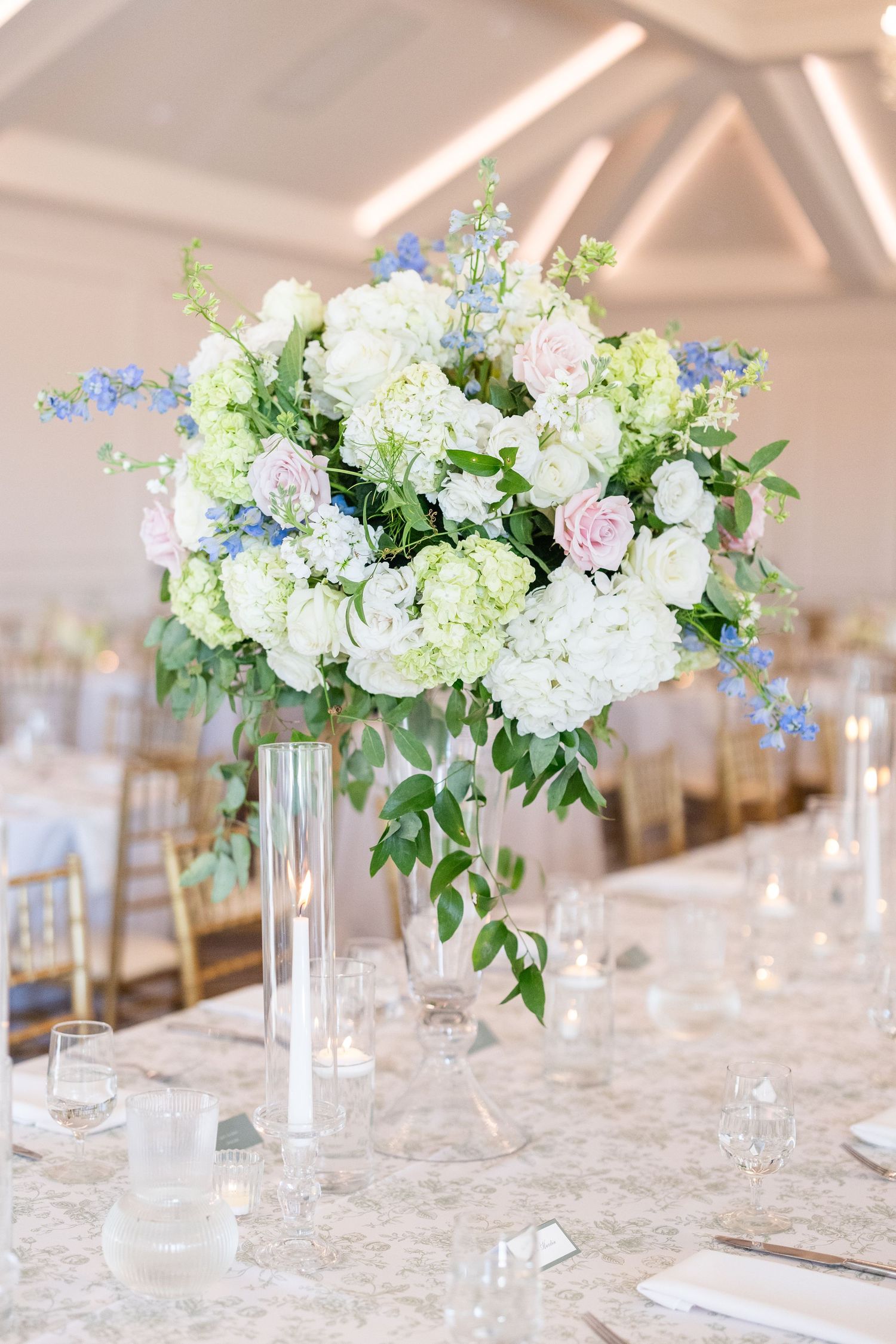 Tall elegant centerpiece with white hydrangeas, pink roses, and blue delphinium on a reception dinner table.