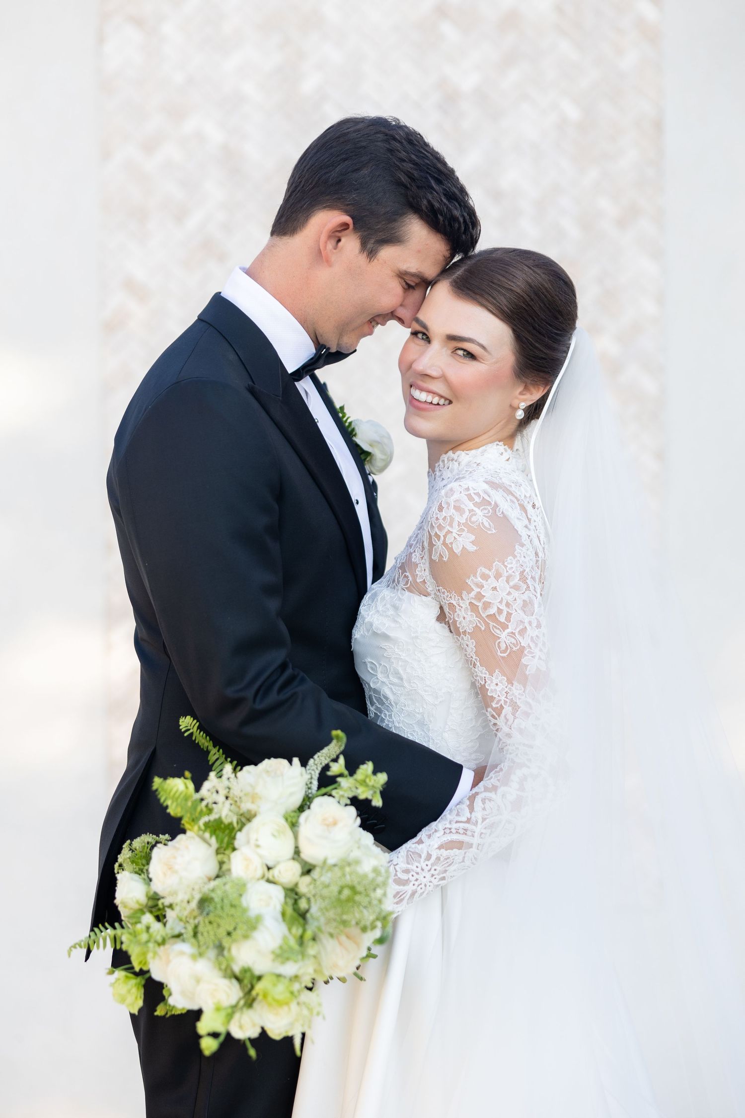Bride in lace long-sleeve gown smiles as groom holds her close before a white floral backdrop.