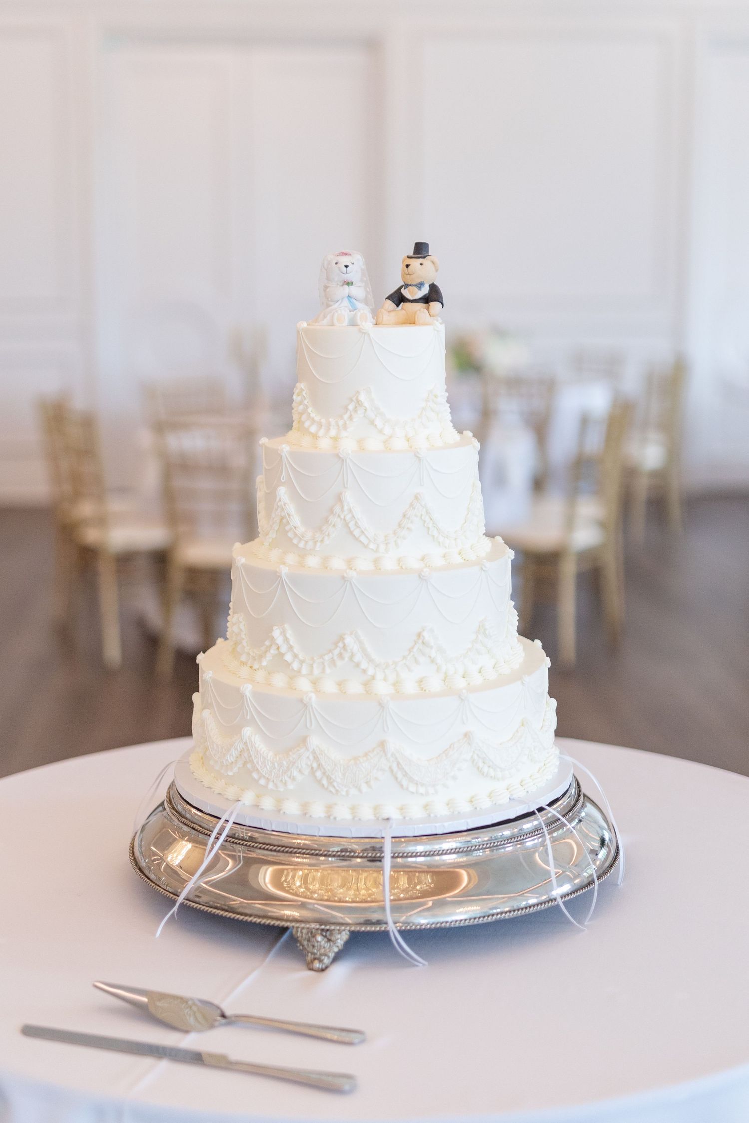 Four-tier white wedding cake with pearl and drape piping details and teddy bear bride and groom cake toppers.
