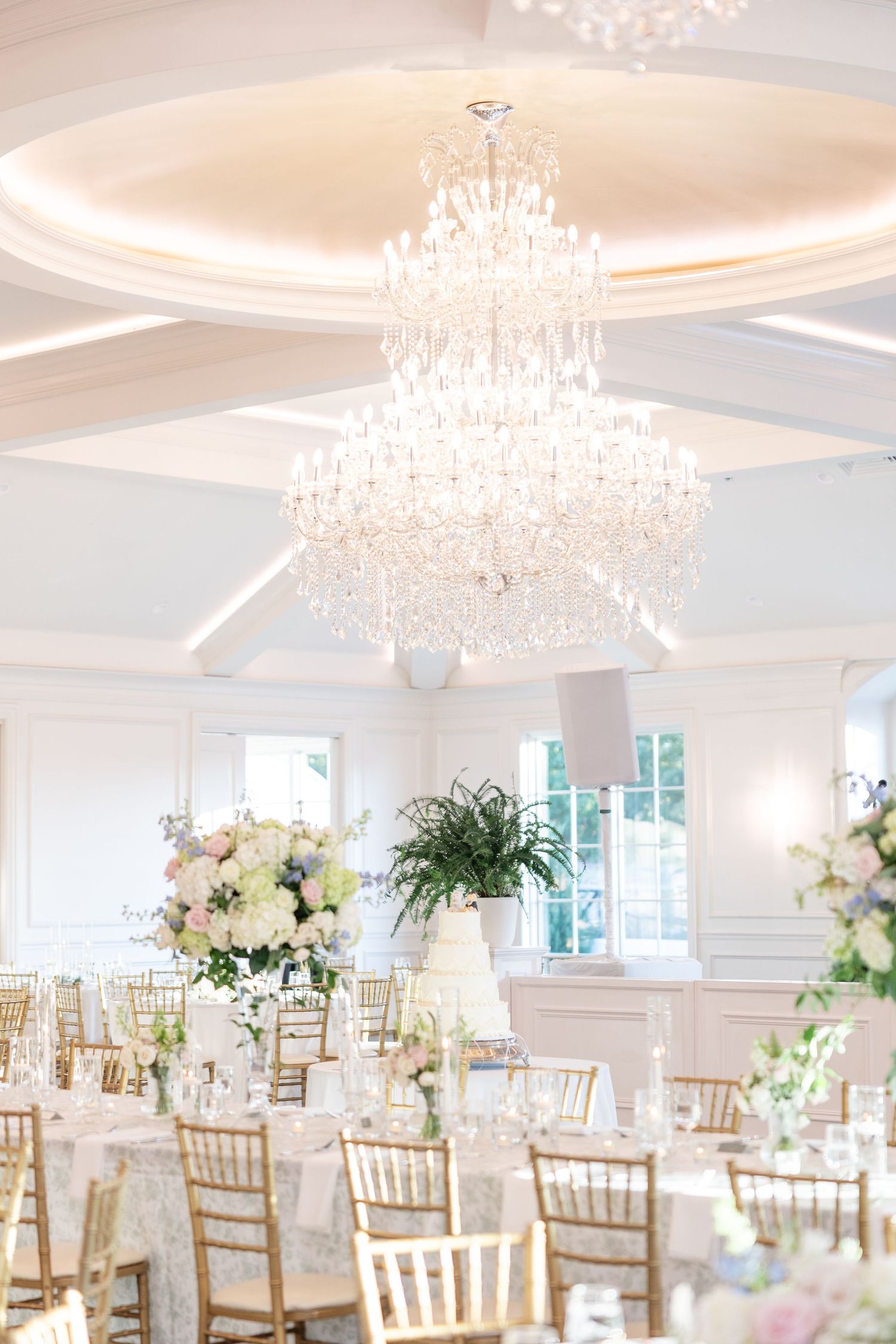 Luxurious white wedding reception ballroom with grand crystal chandelier, gold chiavari chairs, and floral centerpieces.