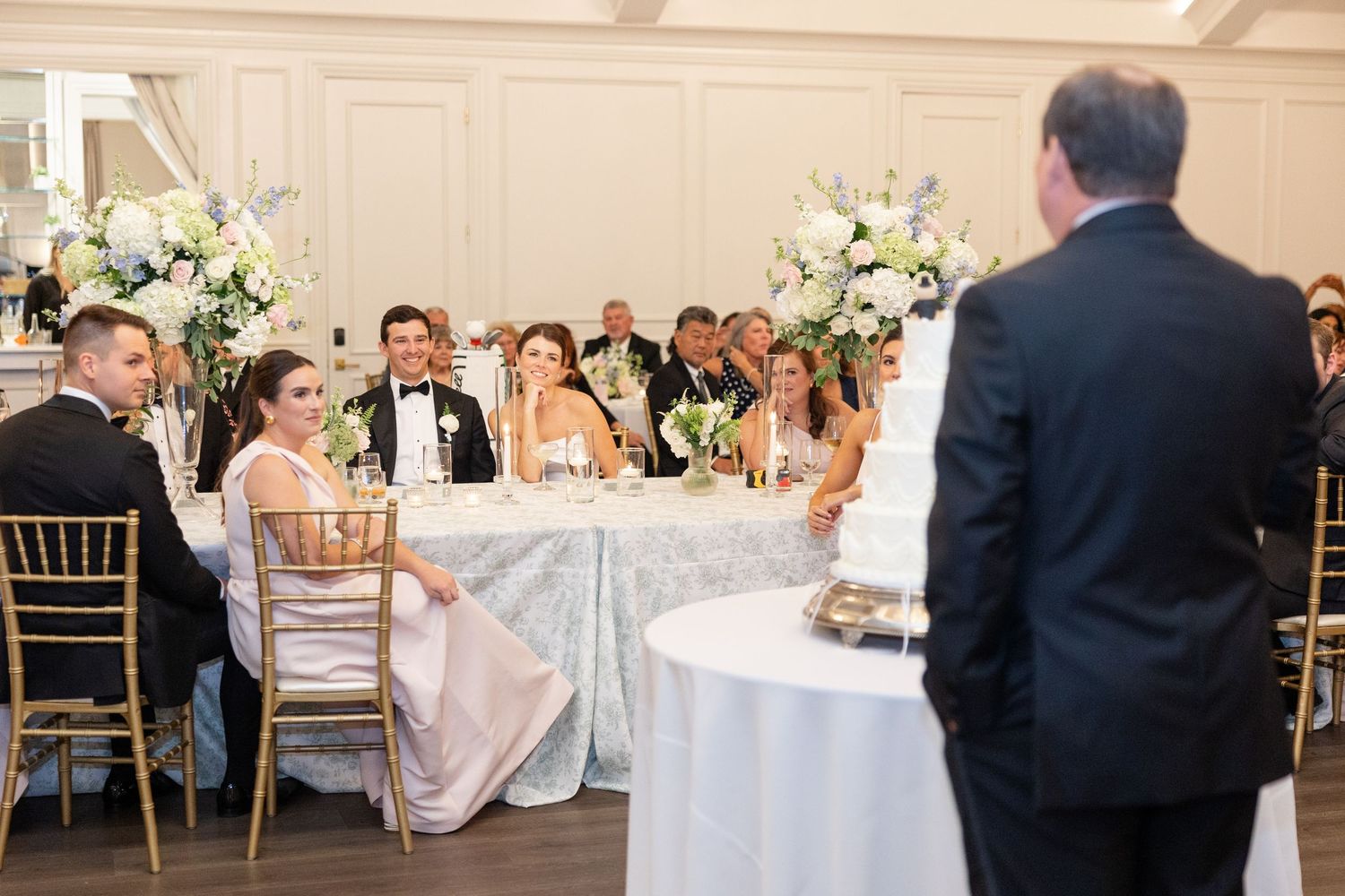 Wedding party seated at head table smiling during reception speeches in elegant ballroom.