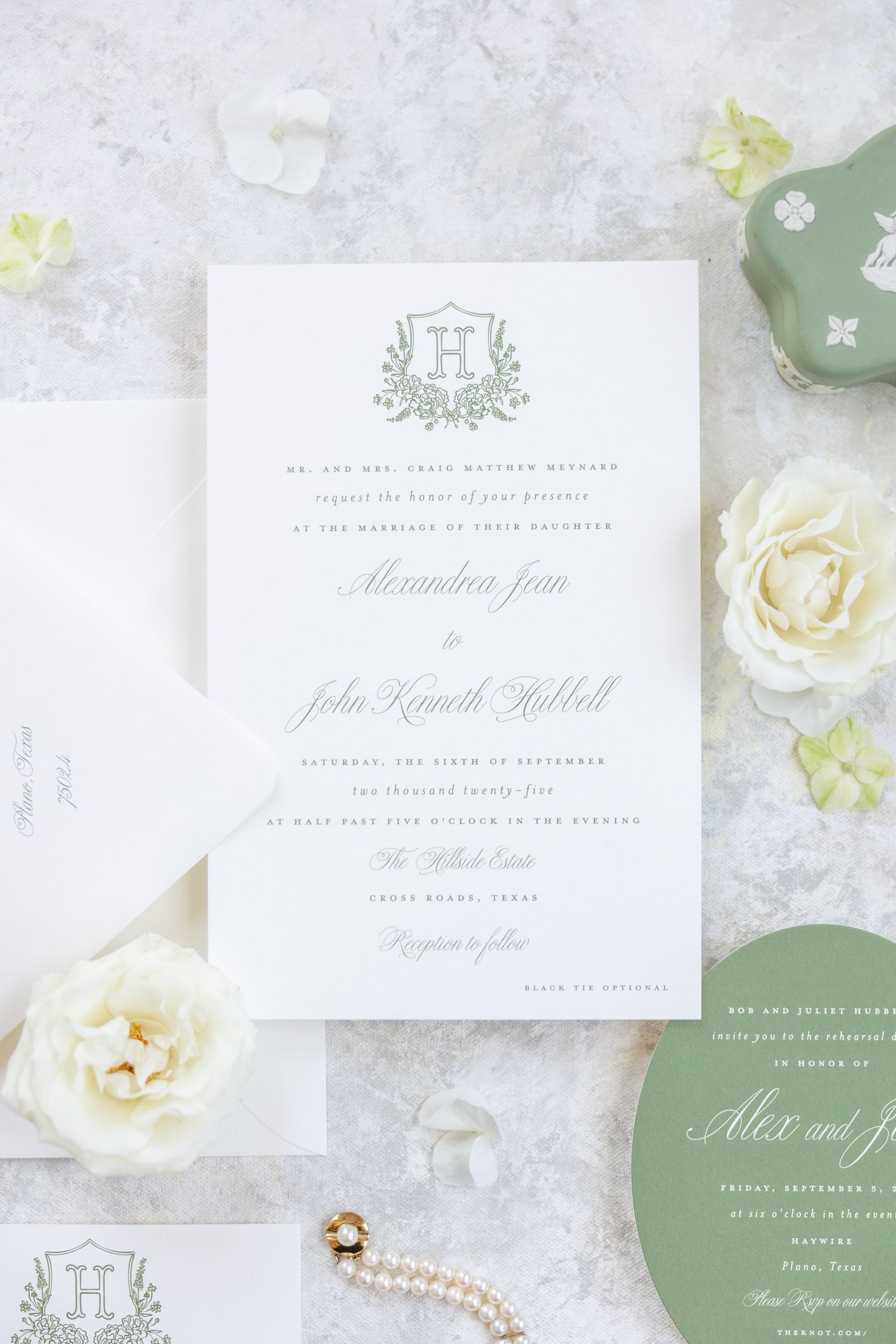 Elegant wedding invitation suite with monogram crest, white roses, and sage green accents on marble background.