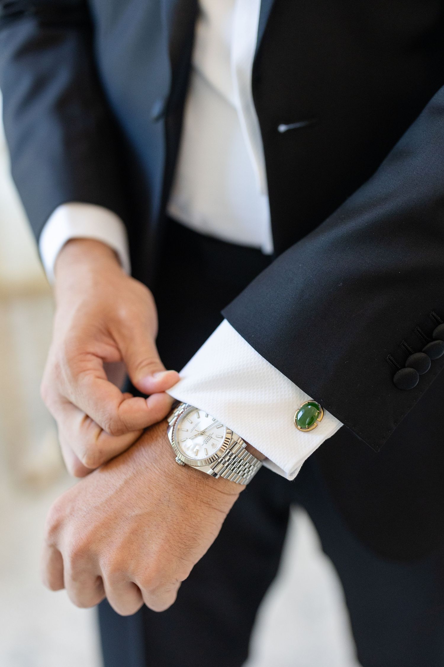 Close-up of groom adjusting silver cufflinks and Rolex watch on tuxedo shirt sleeve.