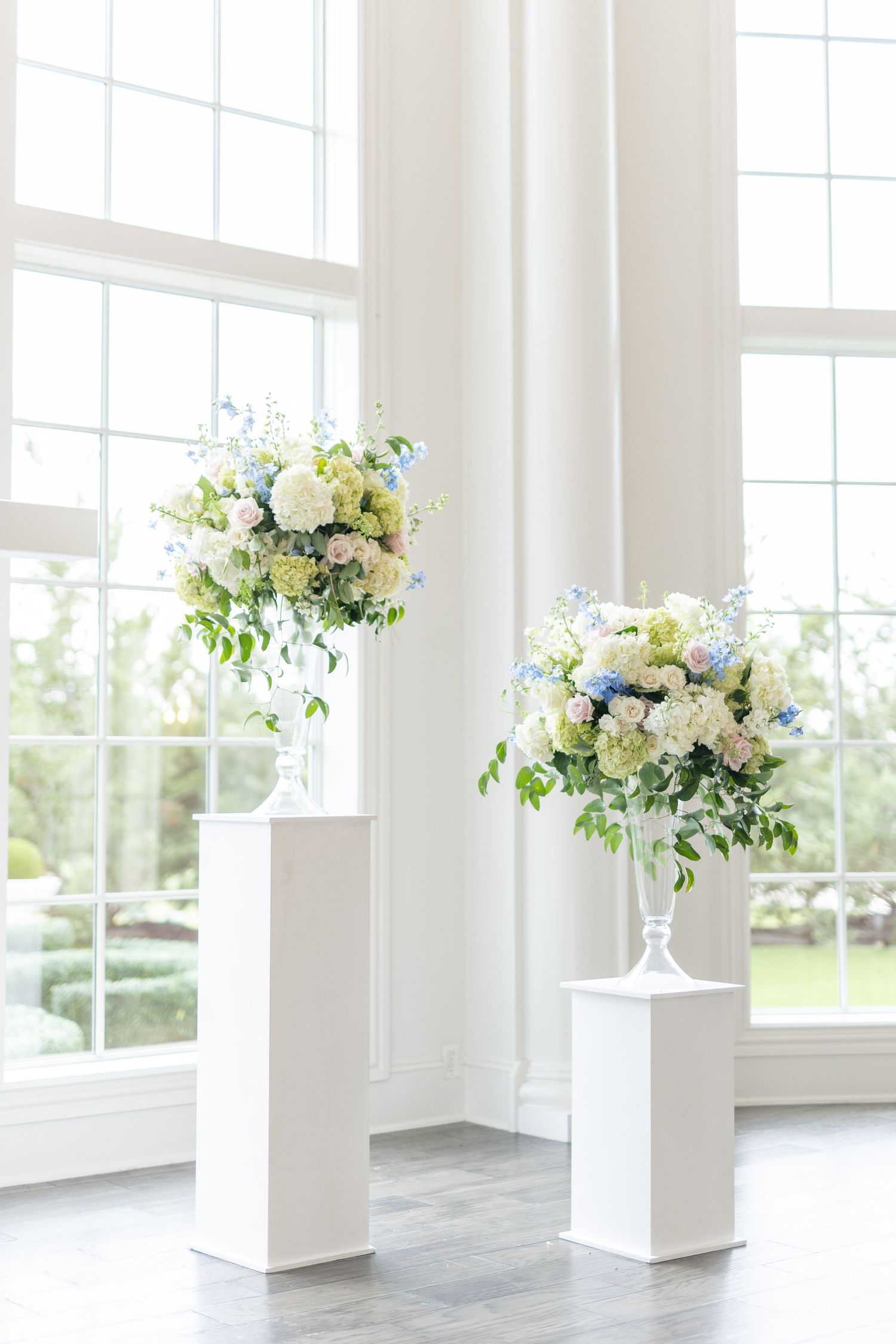 Two elegant floral arrangements of white and blue flowers on white pedestals before tall windows.