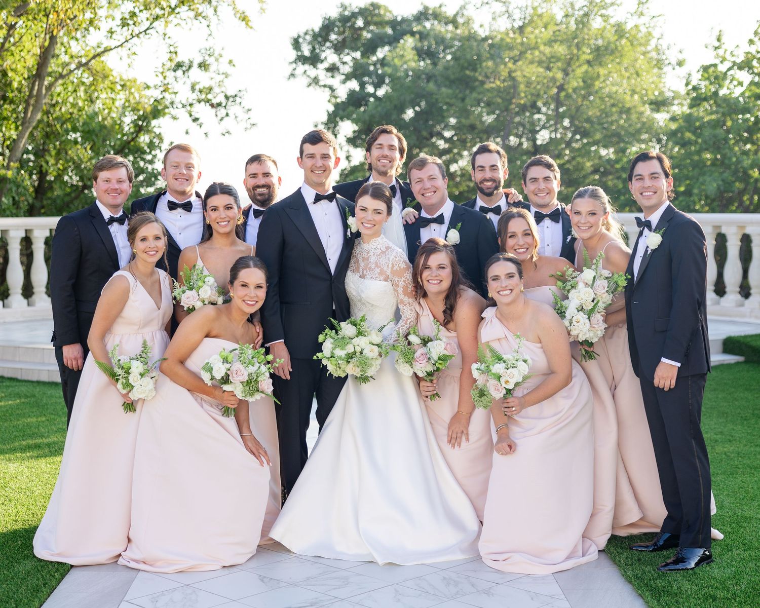 Wedding party poses outdoors, bridesmaids in blush gowns with white bouquets, groomsmen in black tuxedos.