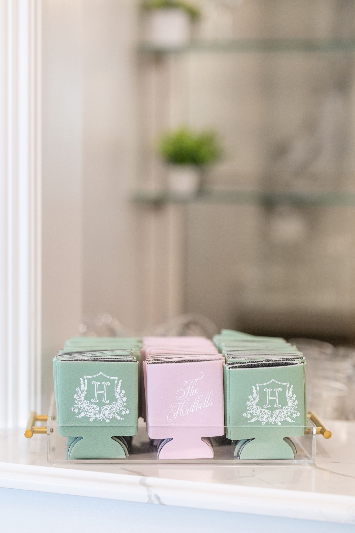Mint green and pink monogrammed koozies with floral crest design displayed in acrylic tray as wedding favors.