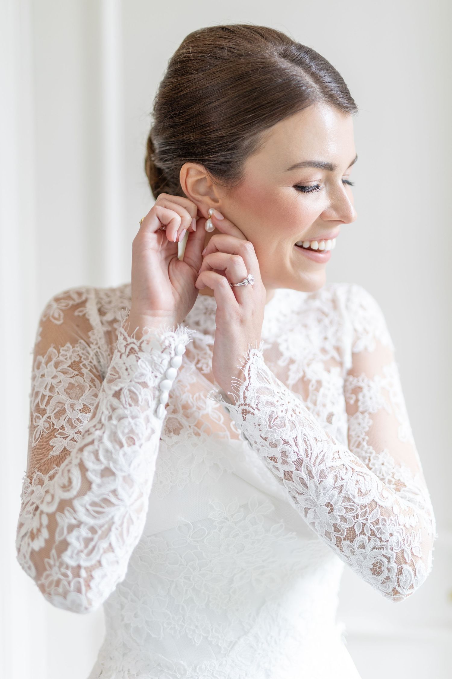 Smiling bride in long sleeve lace wedding dress putting on earrings while getting ready.