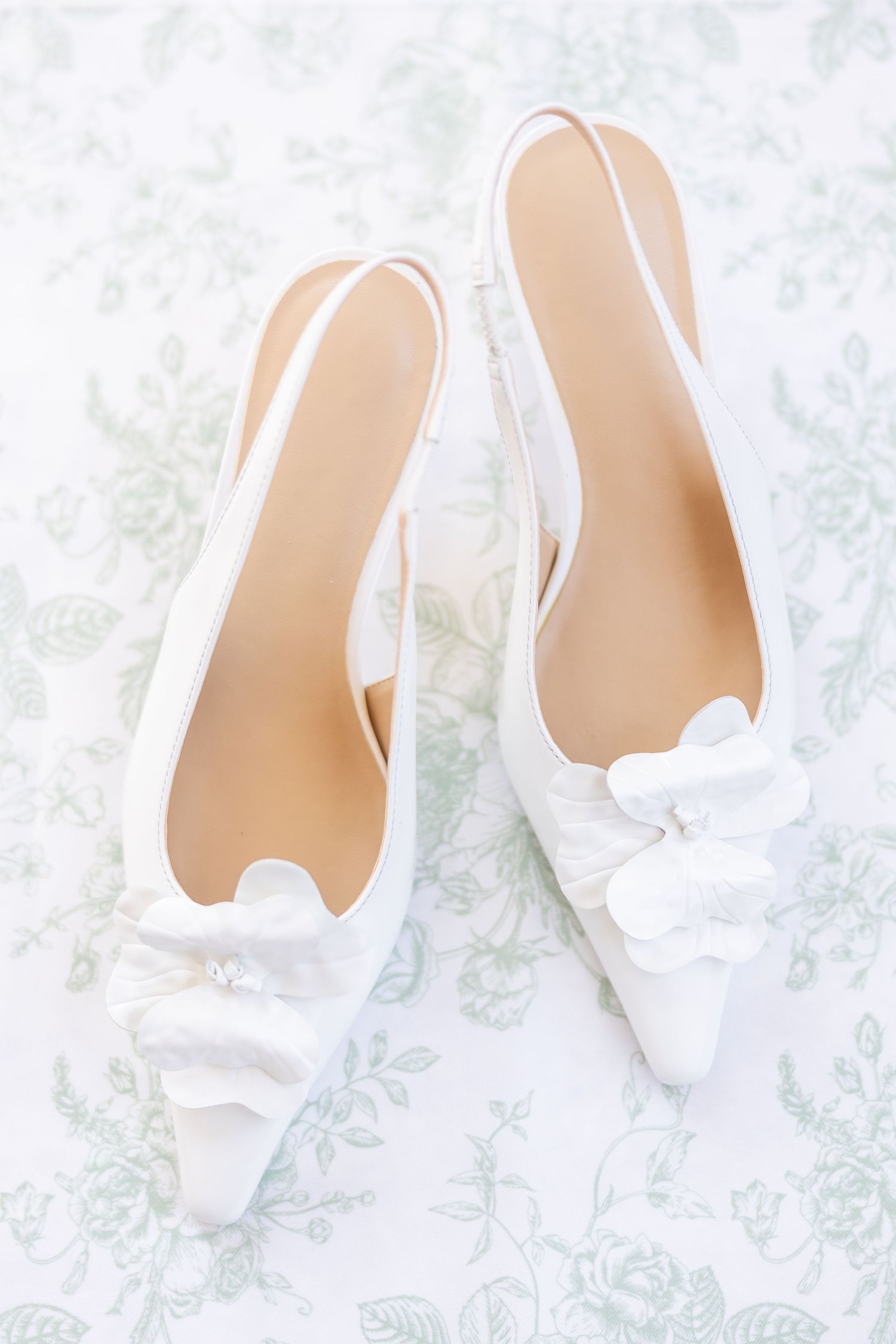 White satin slingback wedding shoes with floral bow detail on soft green floral fabric background.