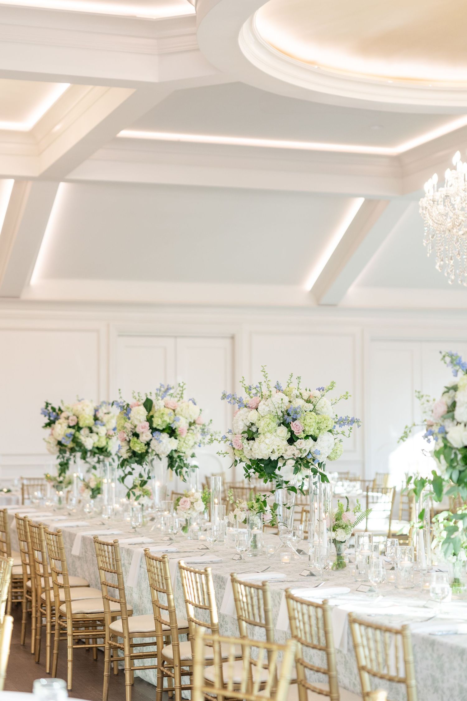 Long reception table with tall white and blue floral arrangements, gold chiavari chairs, and elegant table settings.
