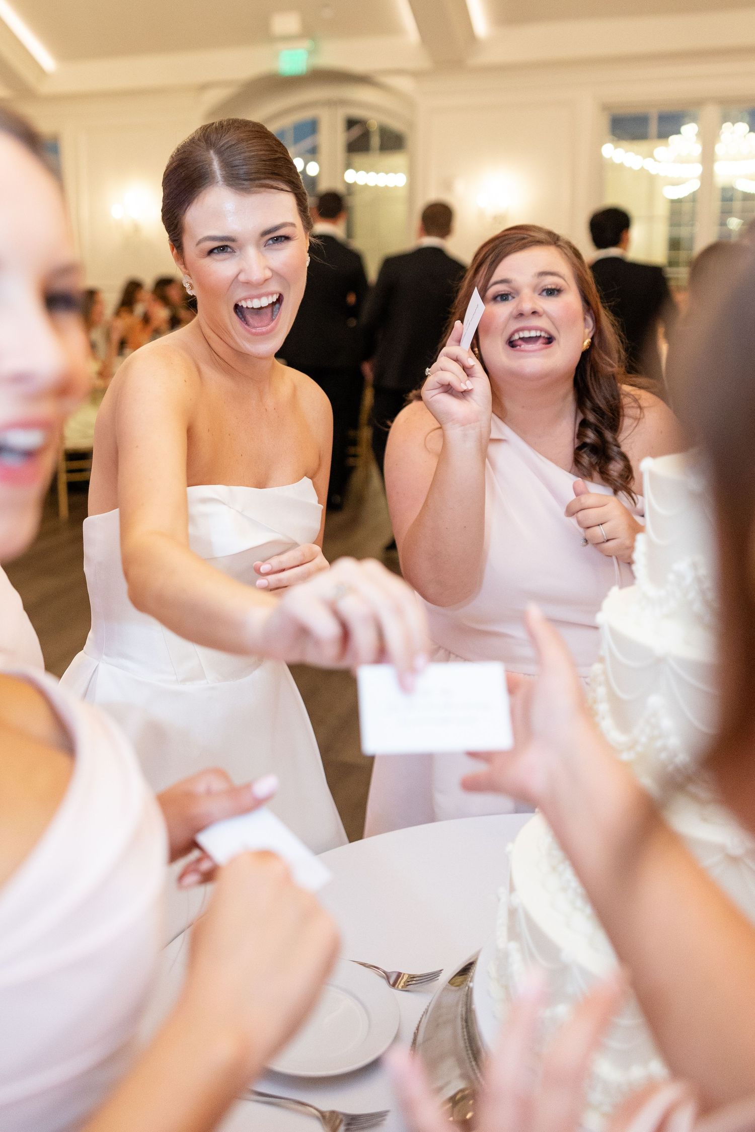 Excited bridesmaids in blush dresses laughing and sharing cards at wedding reception.
