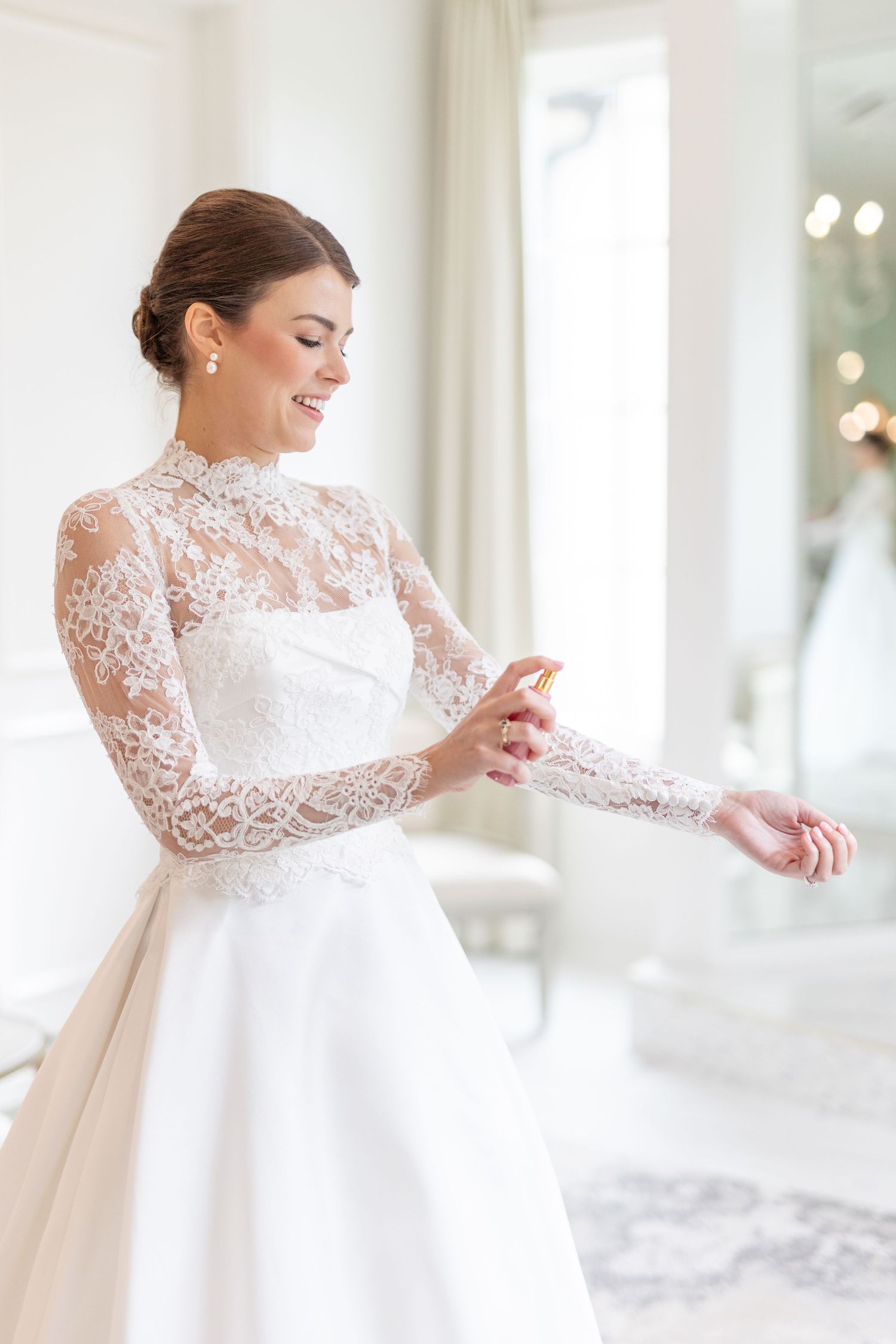 Smiling bride in high-neck lace wedding gown applying perfume to her wrist before ceremony.