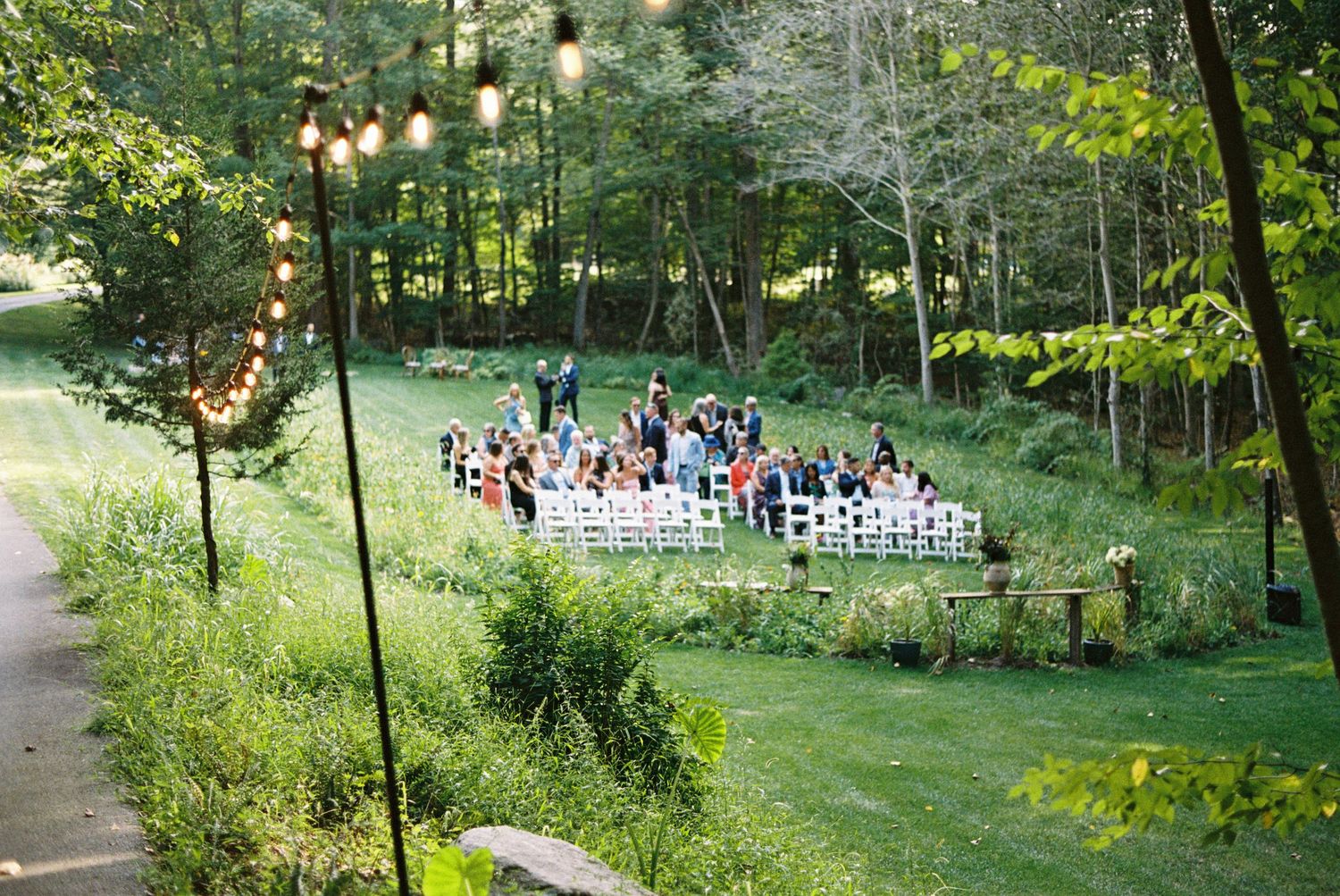 Outdoor wedding ceremony in lush green forest clearing with white chairs and string lights.